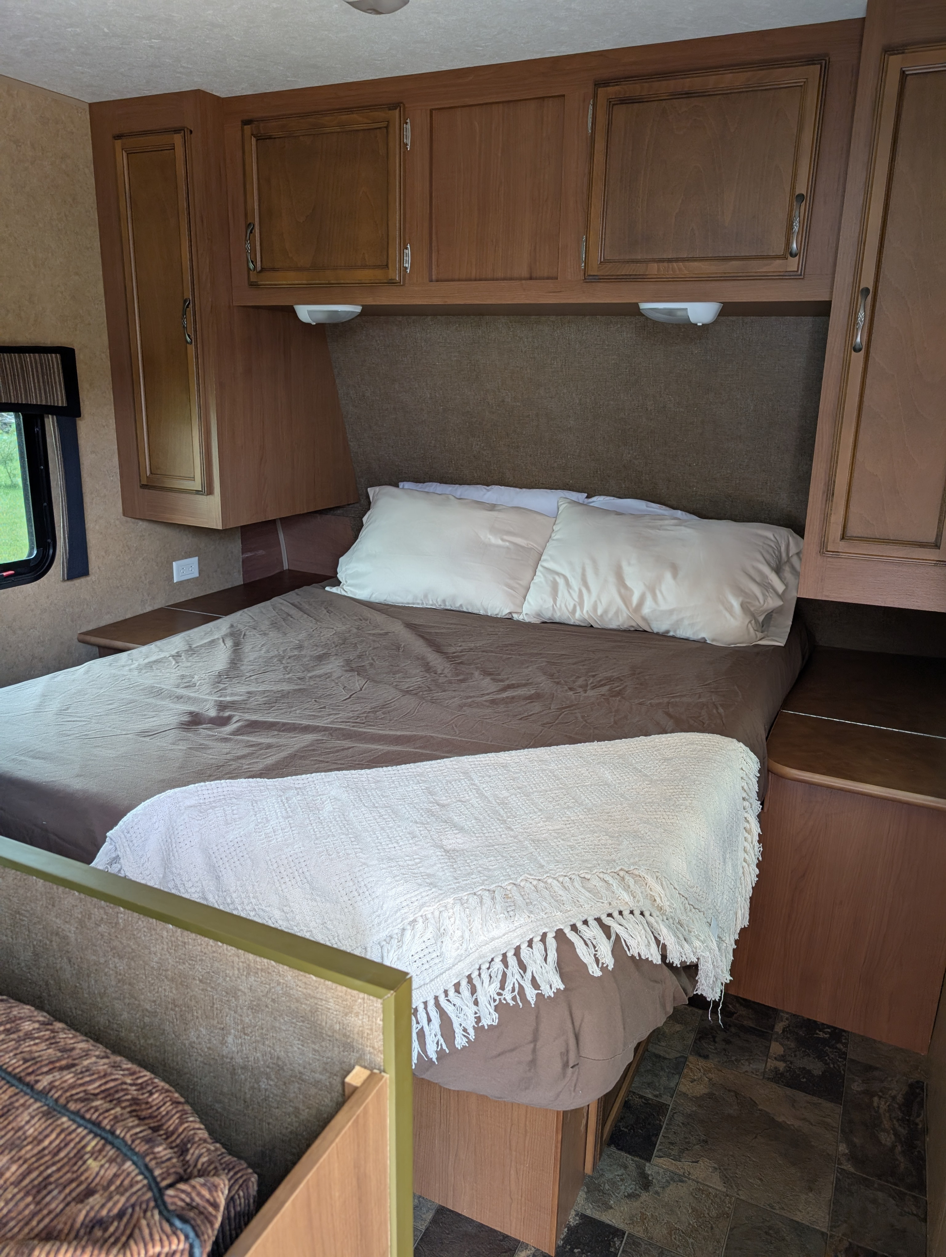 Inside an RV rental, this bedroom features a bed with brown sheets, white pillows and a cream throw, wood cabinets, and a side window.. Starcraft Autumn Ridge 2013