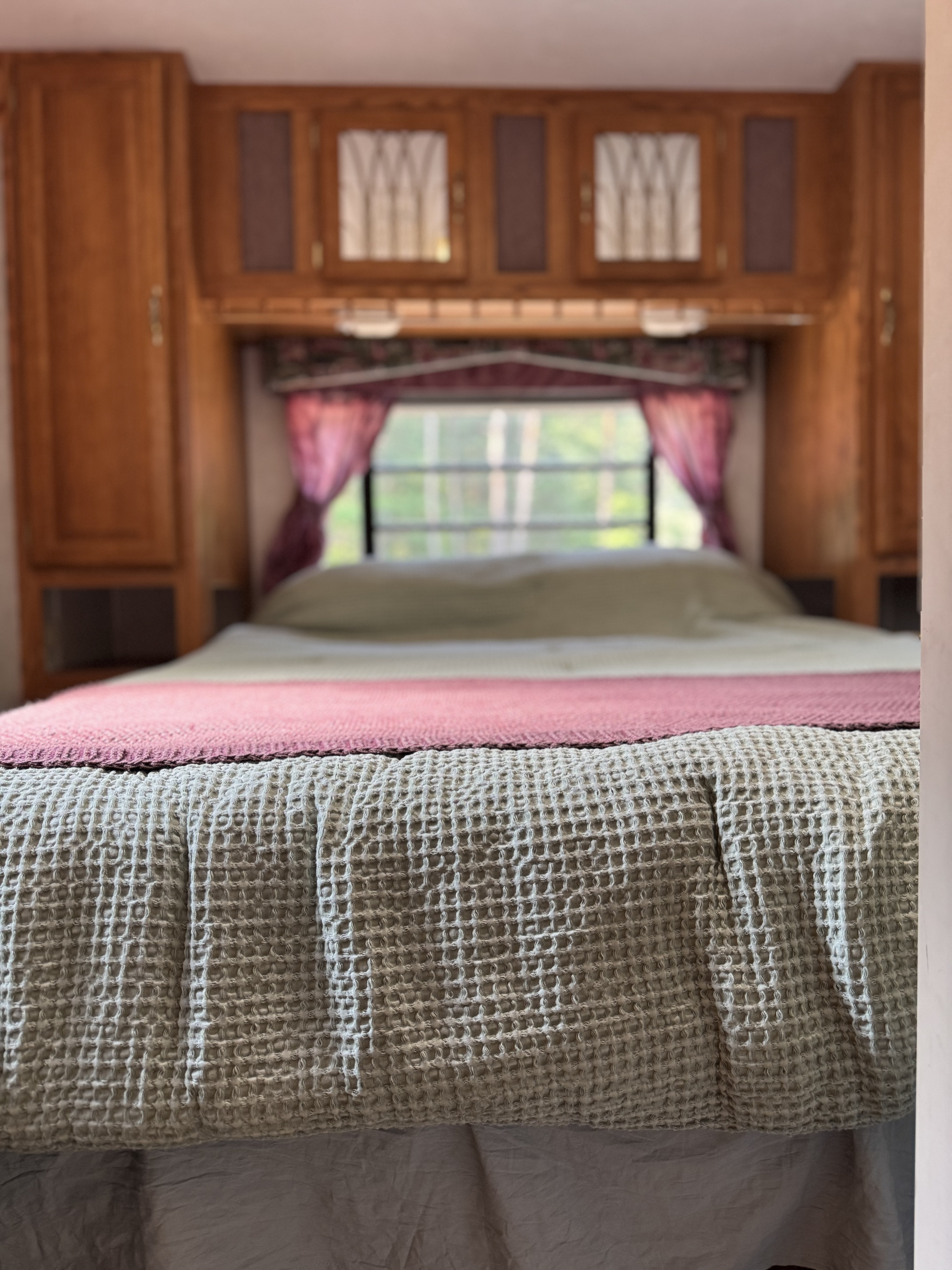 Experience comfort in this RV rental bedroom: a cozy bed with waffle-knit blanket, wooden storage, and nature views.. Forest River Wildwood 2000