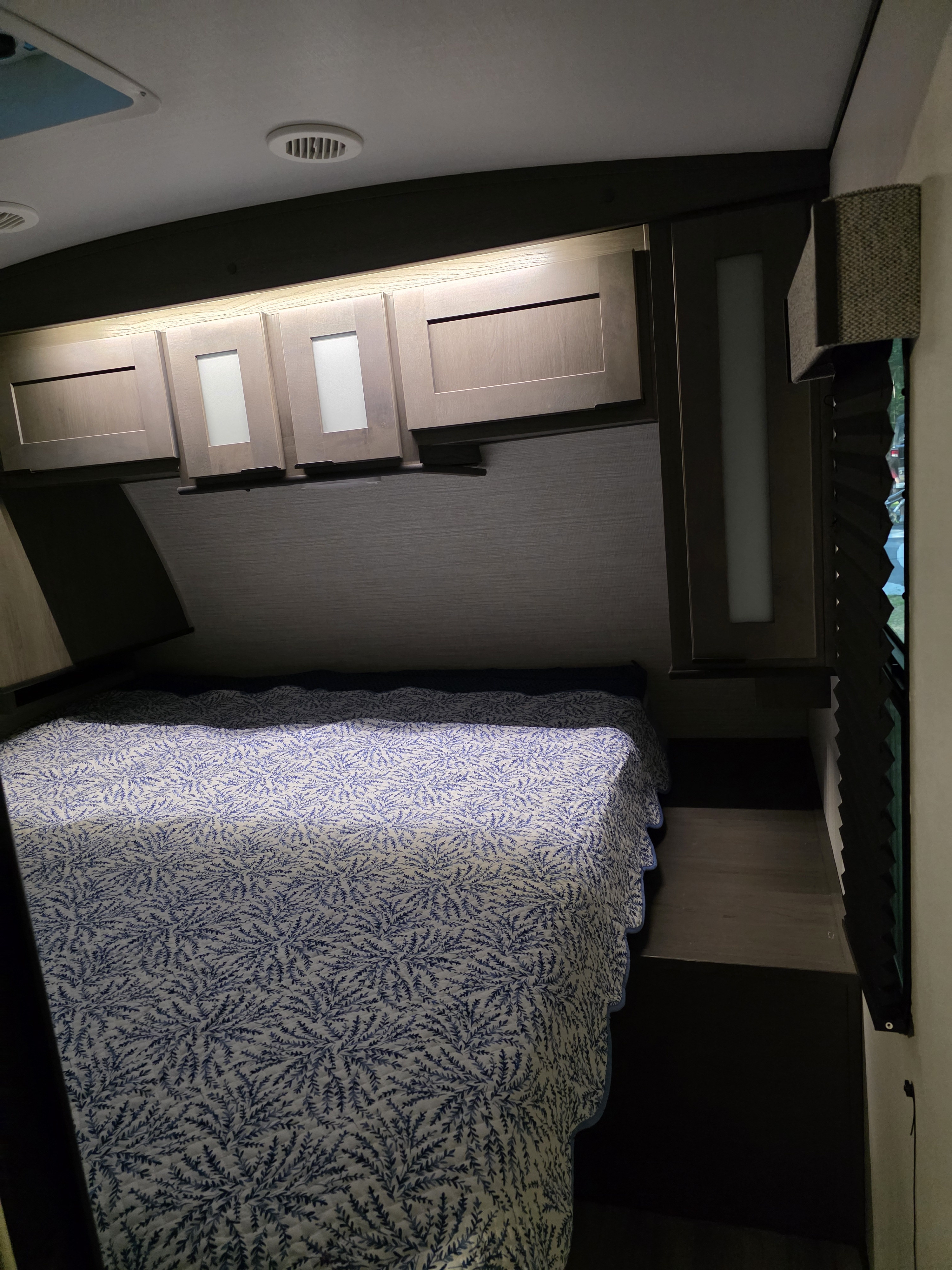 An RV rental bedroom with a bed, blue patterned quilt, overhead cabinets with lighting, and a window for comfortable travel.. Cruiser RV MPG Ultra Lite 2021