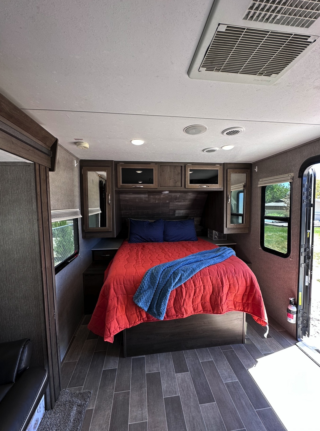 Cozy RV rental bedroom with a comfy red bed, blue pillows, overhead cabinets, and wood-look flooring. Features AC and windows.. Coleman 1805RB 2020