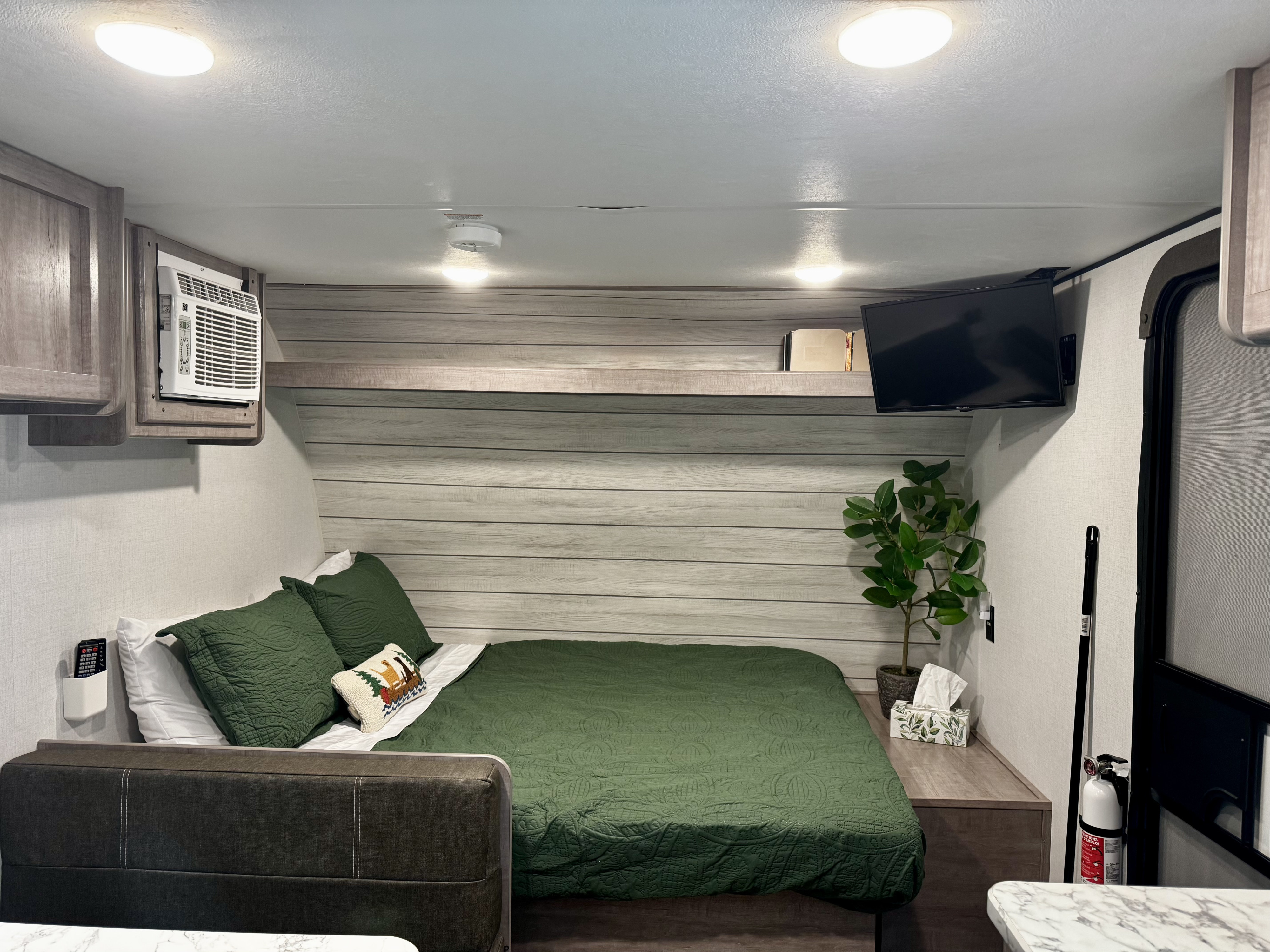 Cozy RV rental bedroom with green bedding, an AC unit, TV, and a small plant, offering comfortable amenities.. Dutchmen Coleman Lantern LT 2023