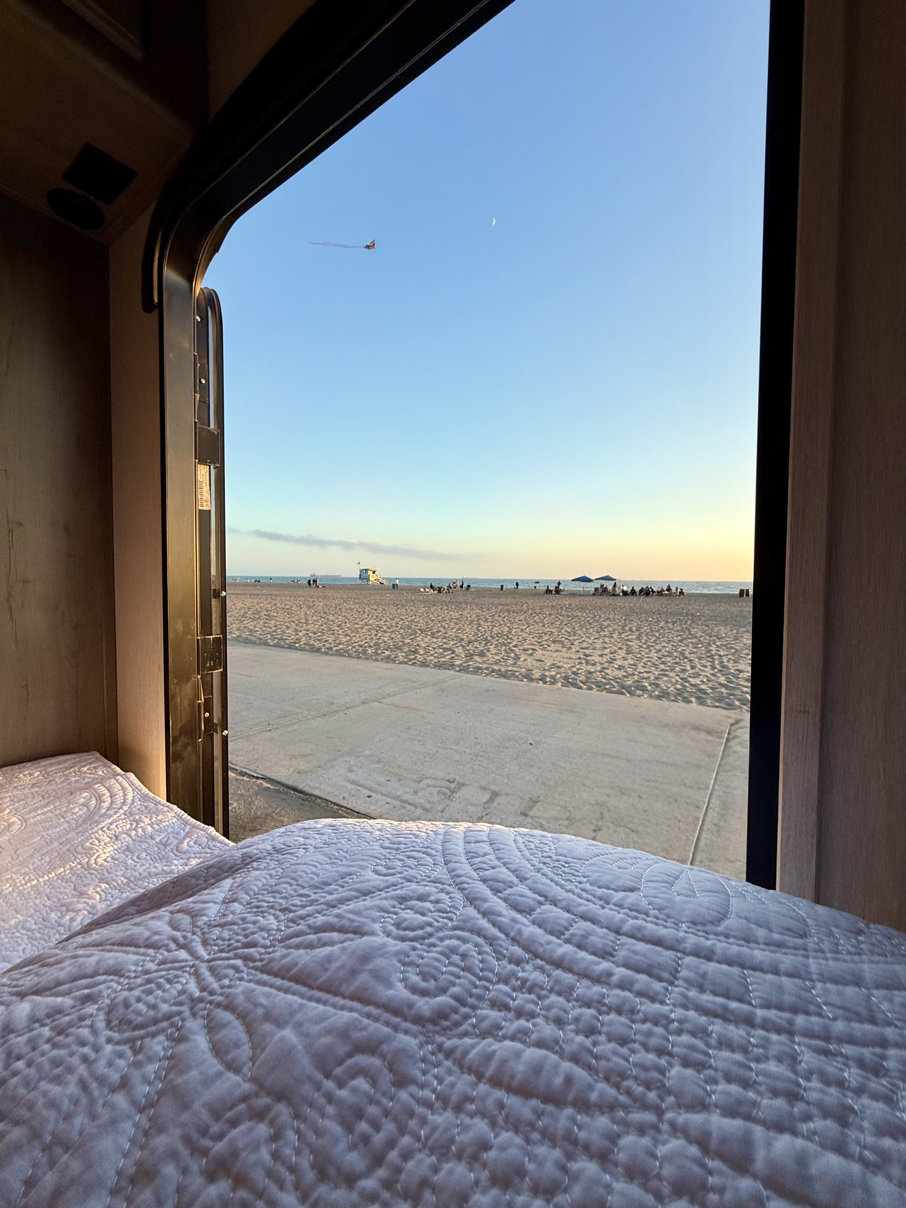 Beachside RV rental view: quilted bed, sandy beach with people, lifeguard tower, and ocean. Perfect sunset escape.. Forest River Coachmen Freelander 2021