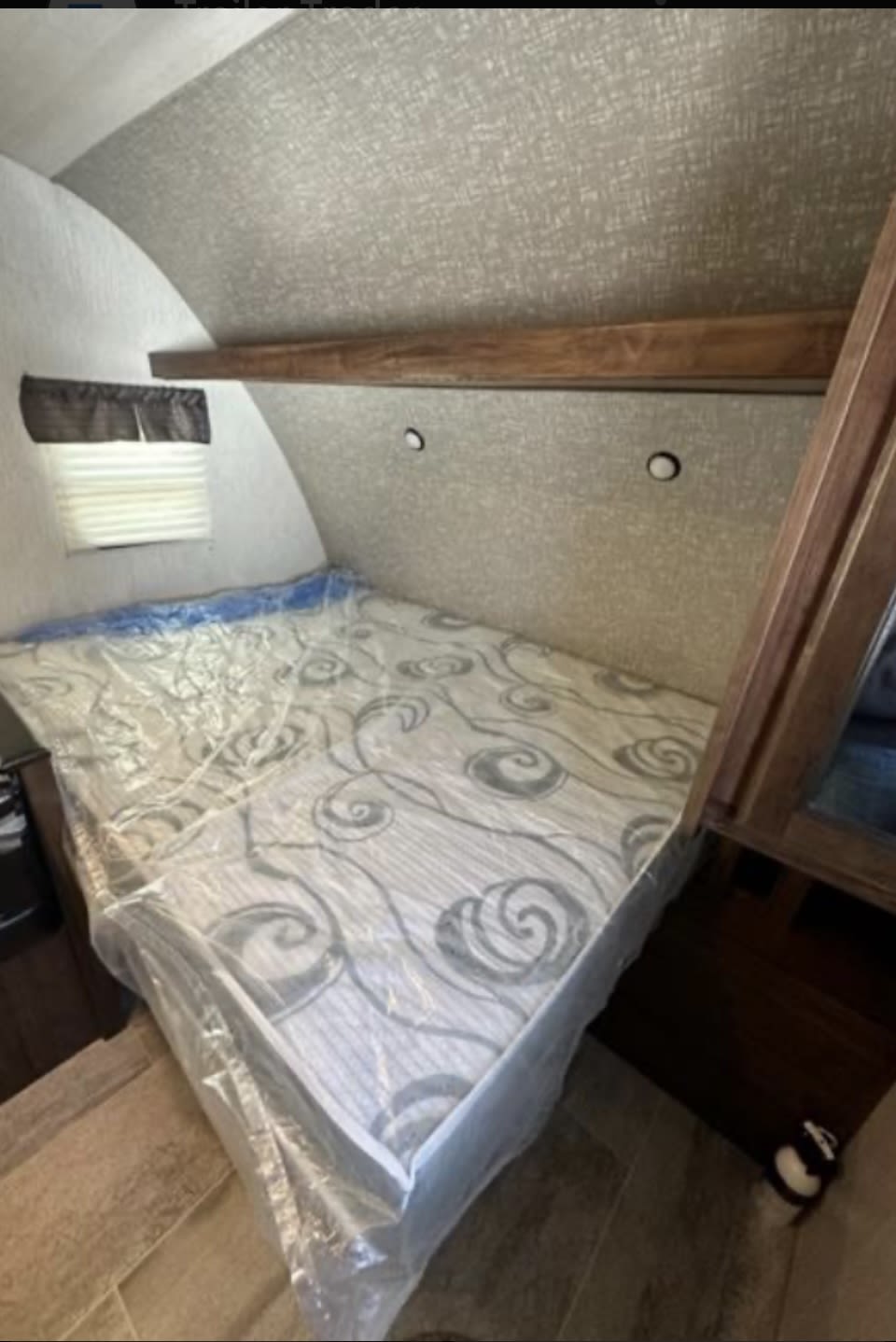Inside an rv rental: A clean bedroom with a plastic-wrapped mattress, small window, and shelf, ideal for comfortable travel and camping.. Mallard M185 2019