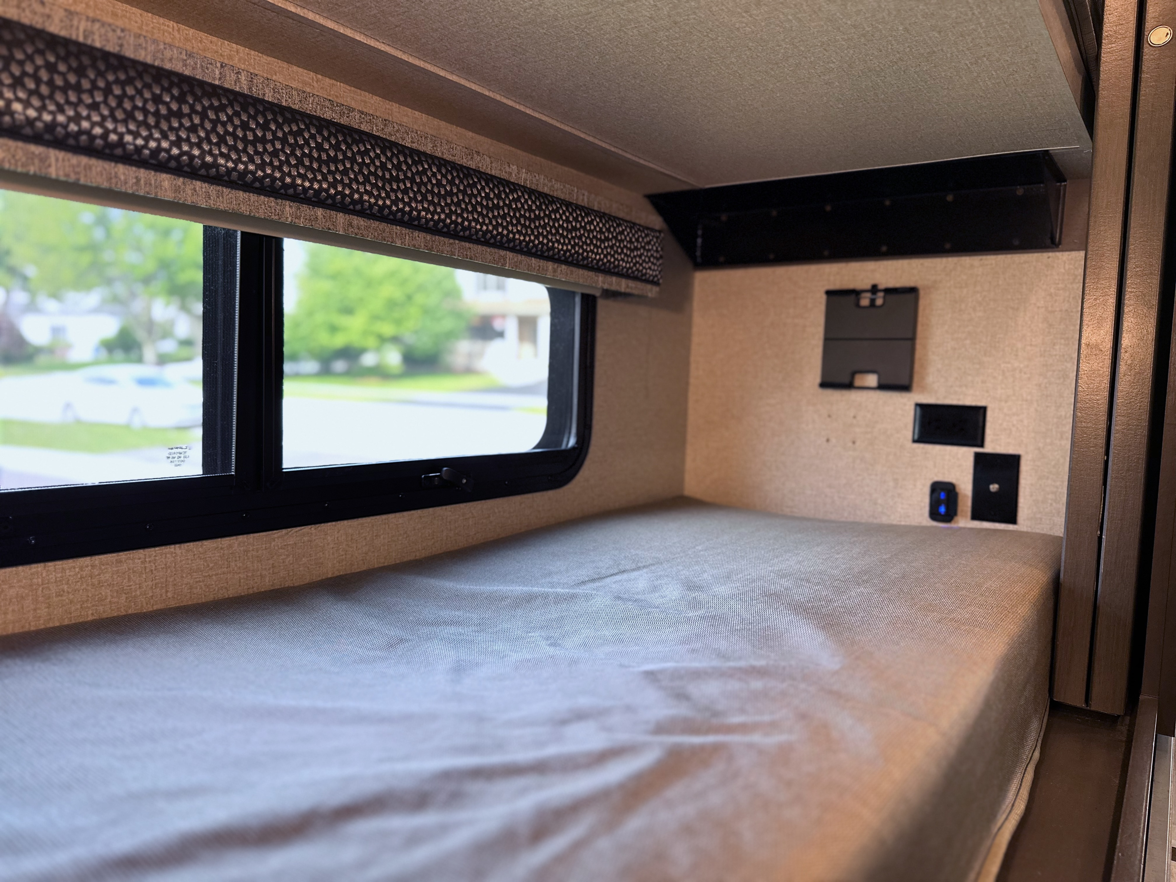 Cozy RV rental bunk bed featuring a window with a green view, integrated power outlets, and stylish valance.. Thor Luminate 2023