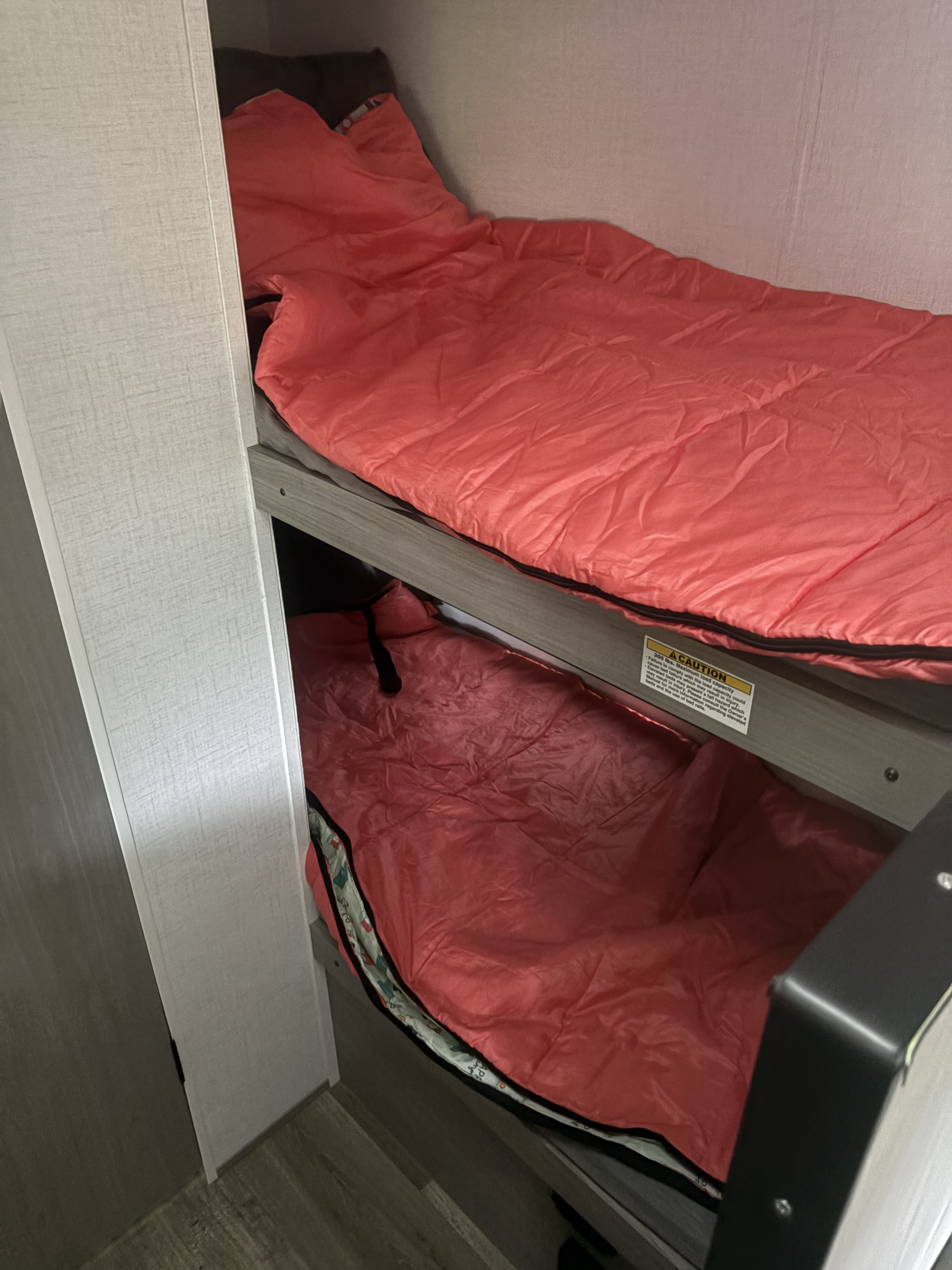 Inside an RV rental: two bunk beds with vibrant red sleeping bags, ready for travelers. Max load 300 lbs per bed caution.. Dutchmen Coleman Latern 2024