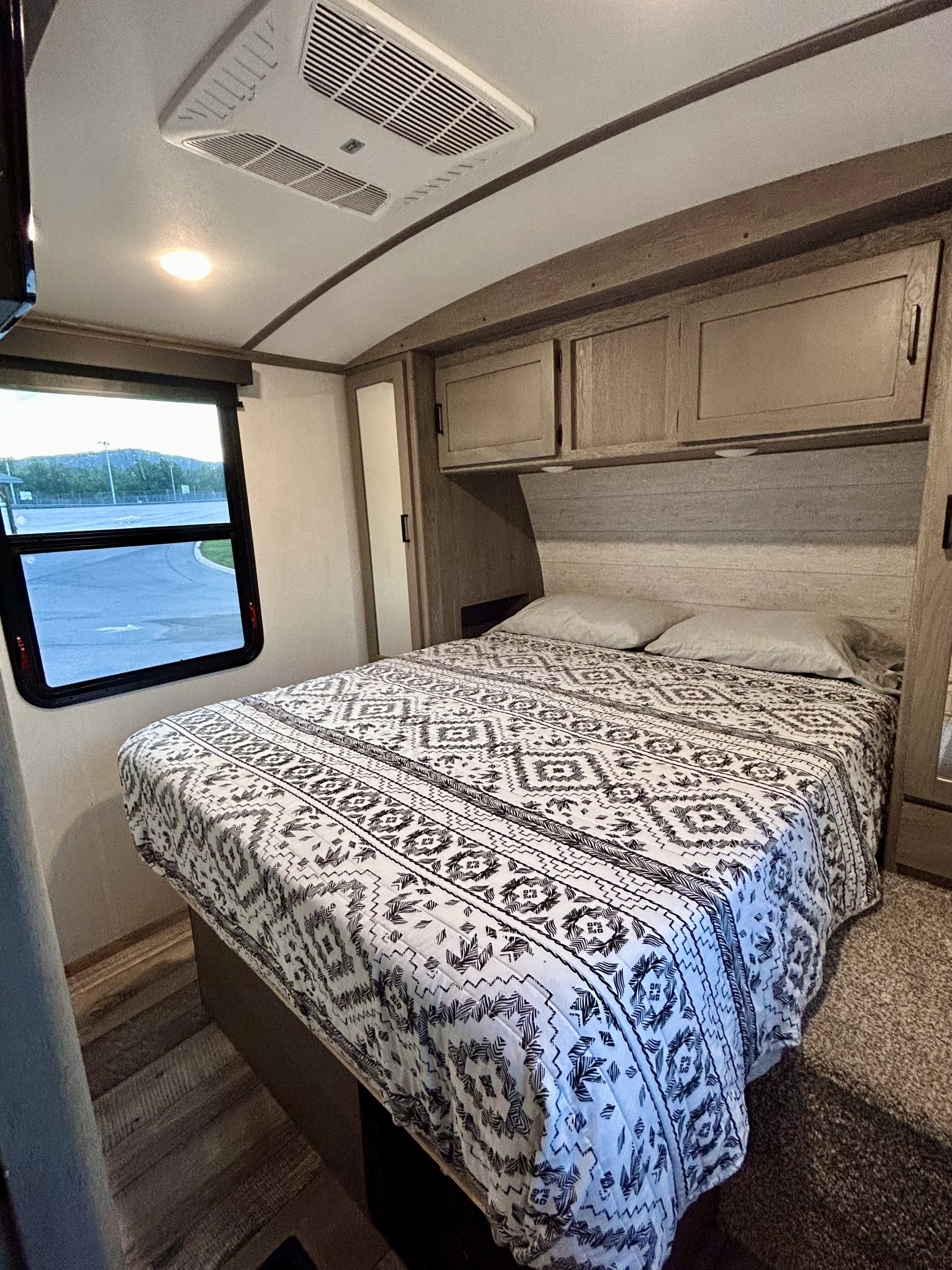 Cozy RV rental bedroom with patterned bedspread, overhead cabinets, and a scenic window view.. Keystone RV Outback 2021