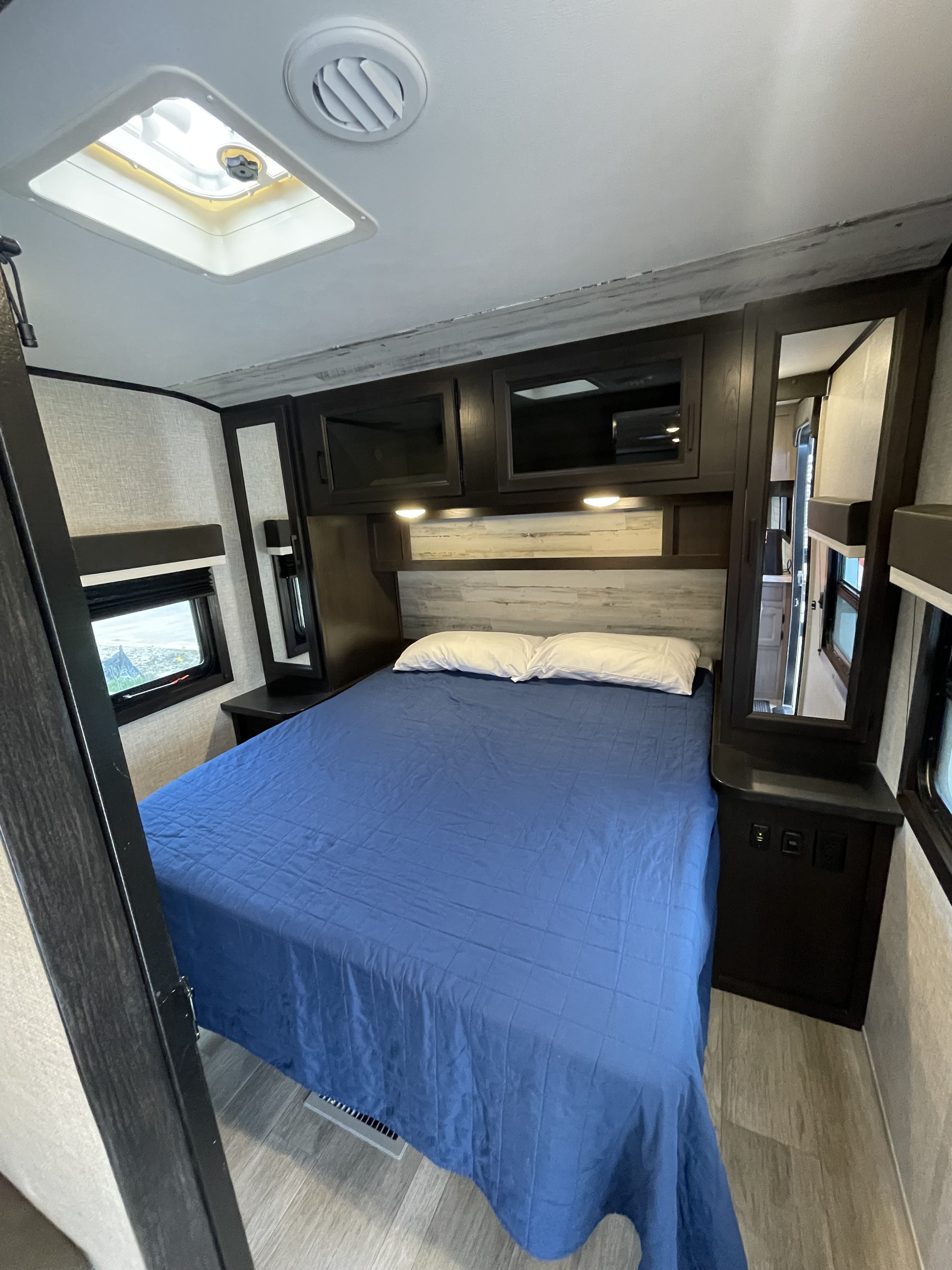 Cozy RV rental bedroom with a blue-comforter bed, white pillows, overhead storage, and light-filled windows.. Jayco Jay Feather 2022