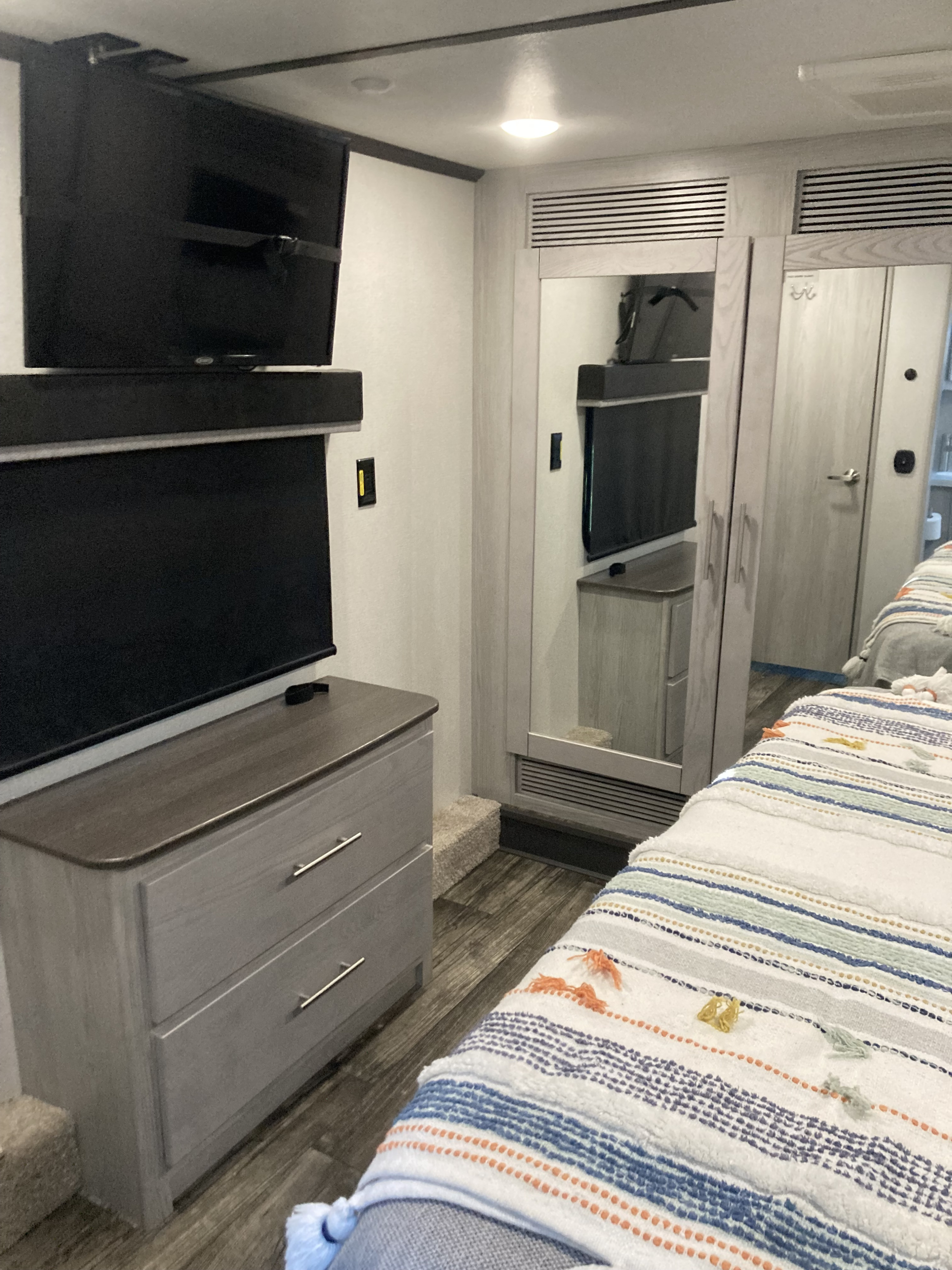 Relax in this RV rental bedroom interior, featuring two TVs (one on dresser, one wall-mounted), a mirrored closet, and a cozy striped bed.. Keystone RV Fuzion 2023
