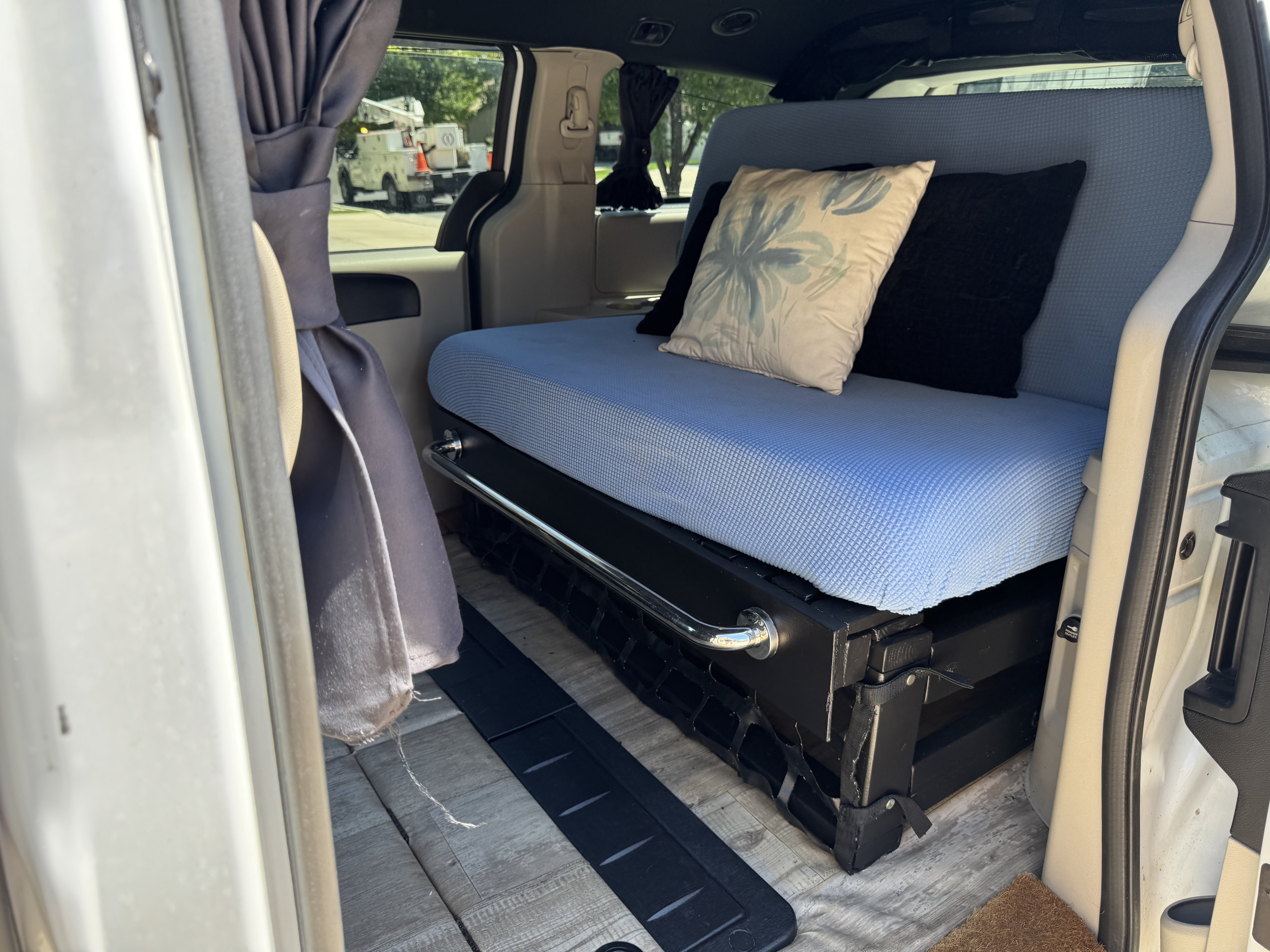 Inside an RV rental, a camper van's custom living space with a blue-covered bed, pillows, and a silver grab bar.. Dodge Grand Caravan 2017