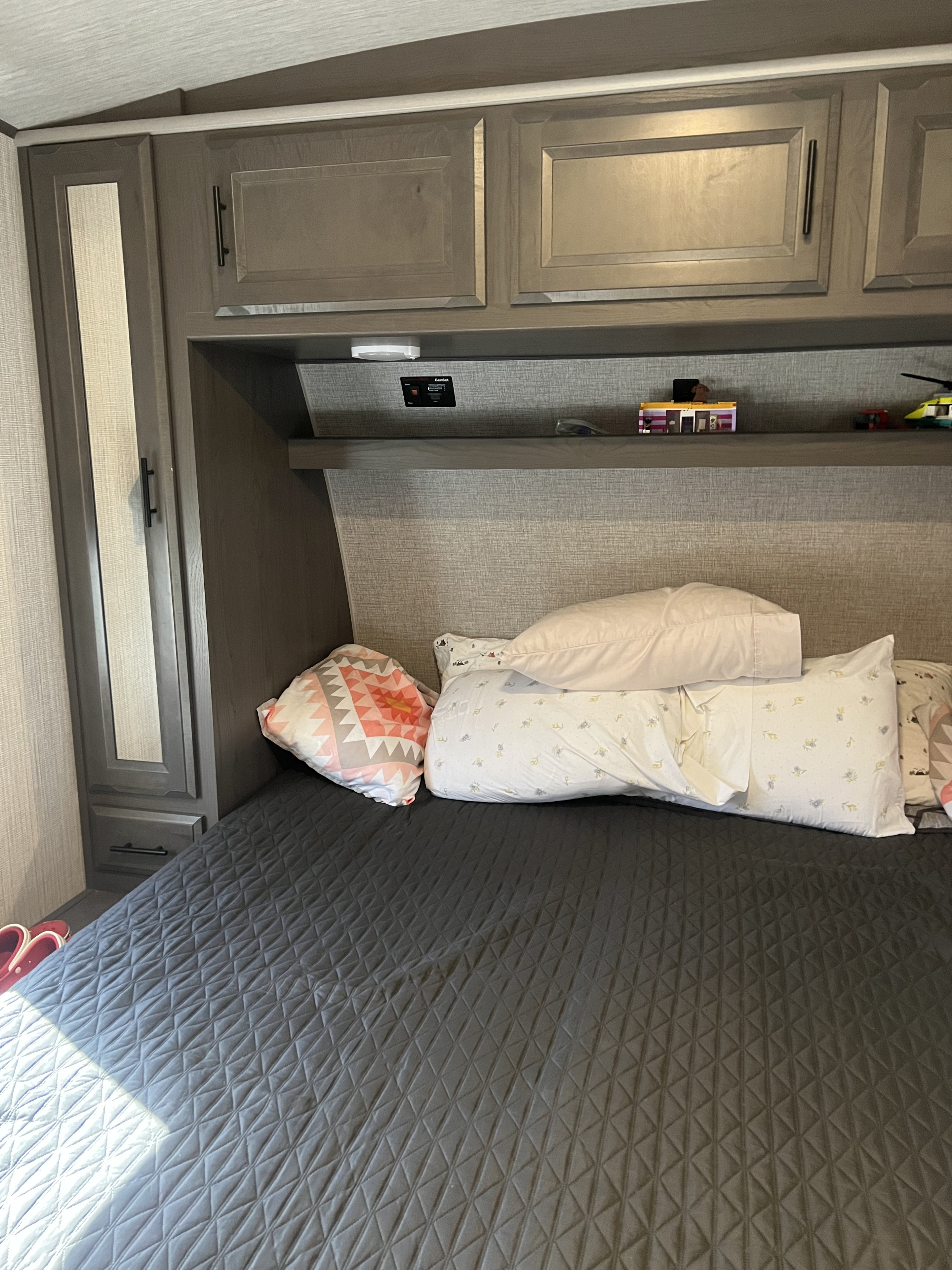 Cozy RV rental bedroom featuring dark gray quilted bed, multiple pillows, and wall-mounted overhead cabinets with a shelf for storage.. Cruiser Rv Corp Stryker 2021