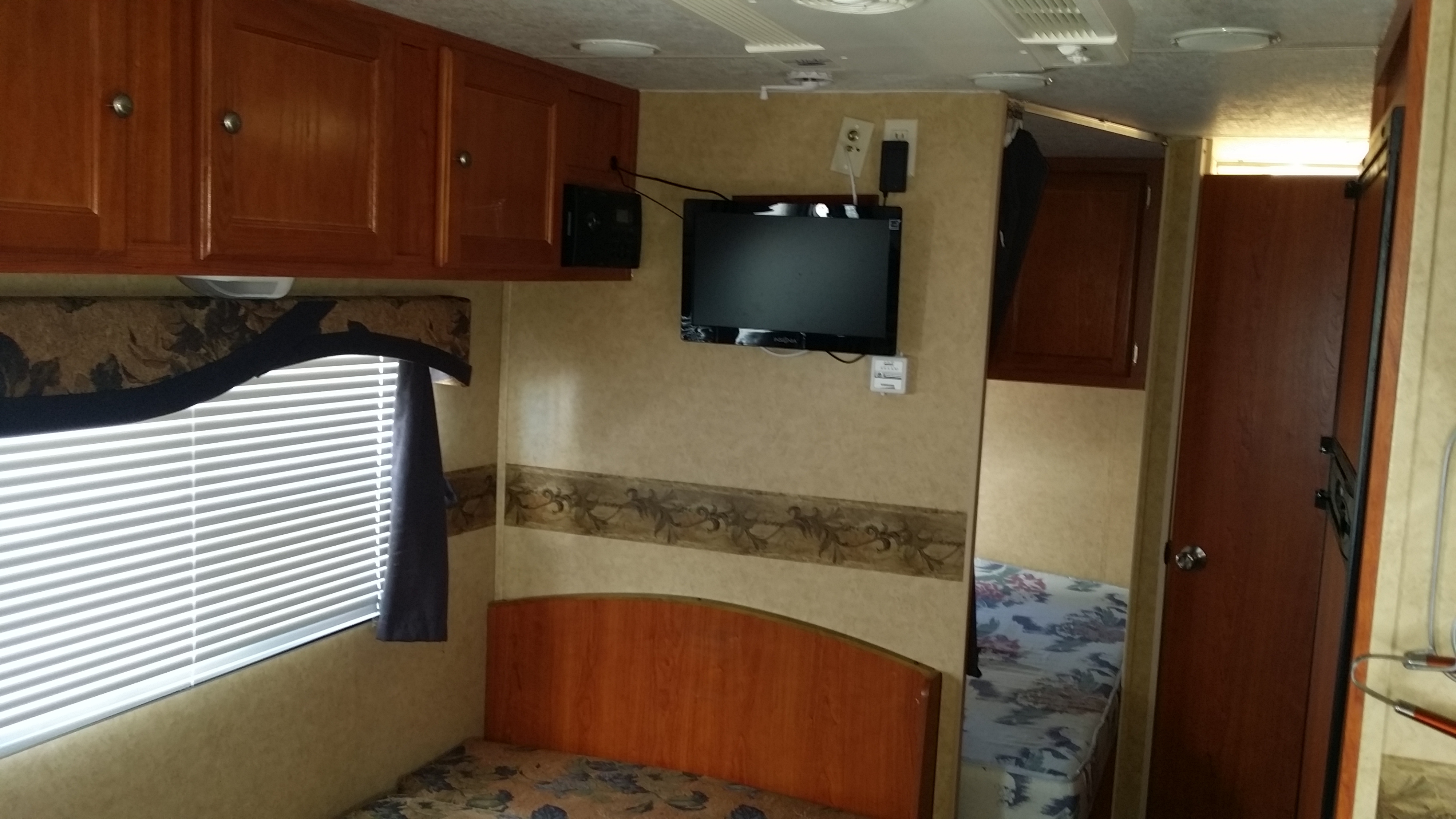 View of an RV rental bedroom with a patterned bed, window with blinds, overhead wood cabinets, and a wall-mounted TV.. R-Vision Super Sport 2010