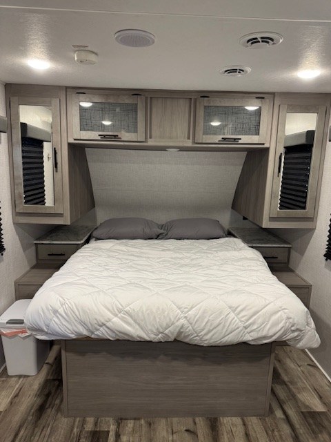 Inside an RV rental: a compact bedroom featuring a queen bed, overhead cabinets, and built-in side storage.. Keystone RV Coleman 1805RB 2022