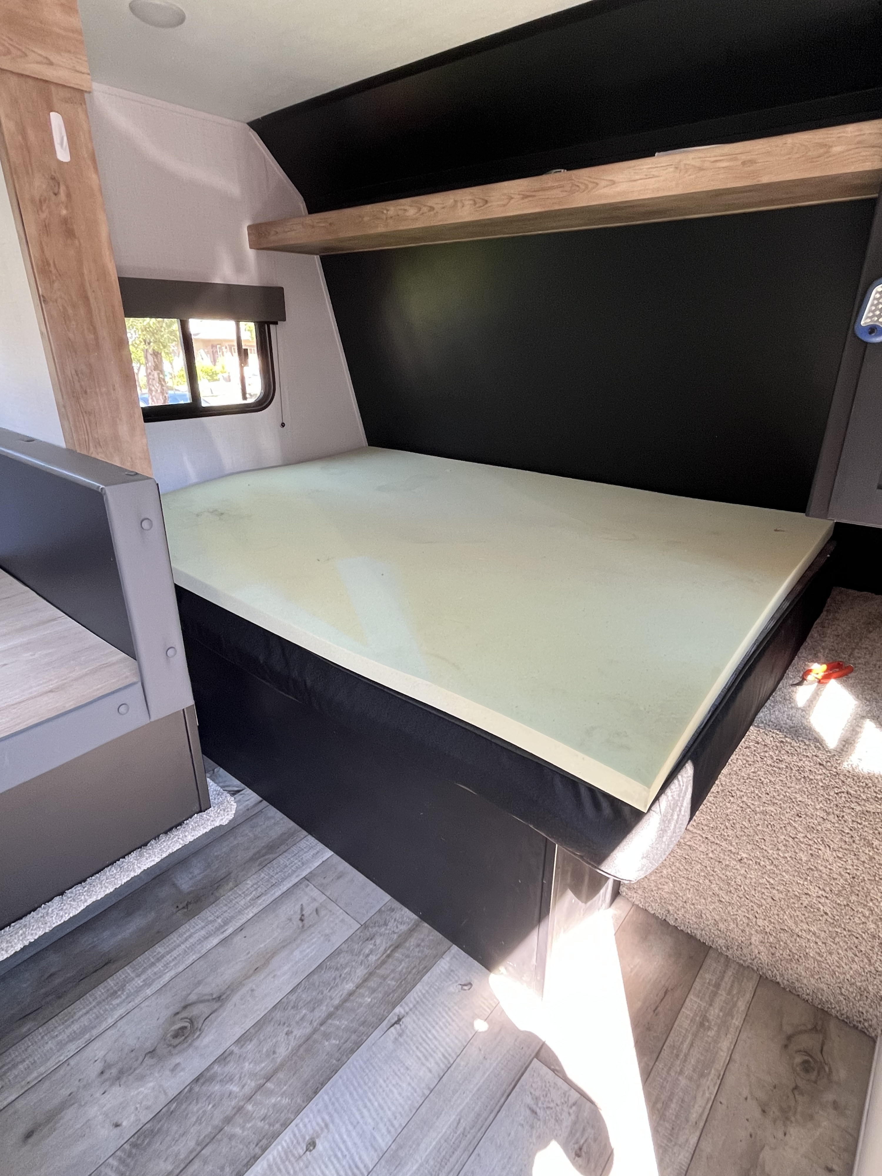 Interior of an RV rental showing a bed with a foam mattress topper, storage shelf, and wood flooring.. Keystone RV Springdale 2022