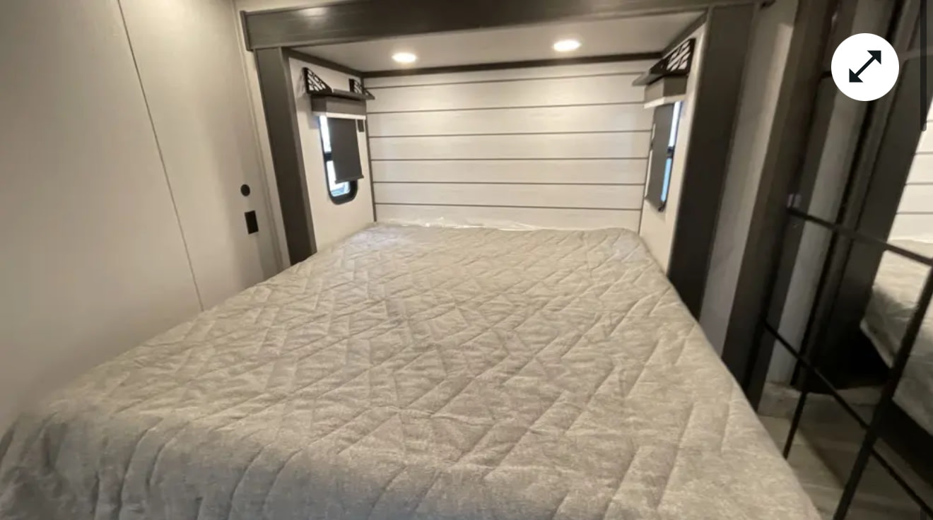 Spacious RV rental bedroom featuring a large bed with a light grey quilted comforter, paneled headboard, and bedside windows.. Keystone Cougar 2025