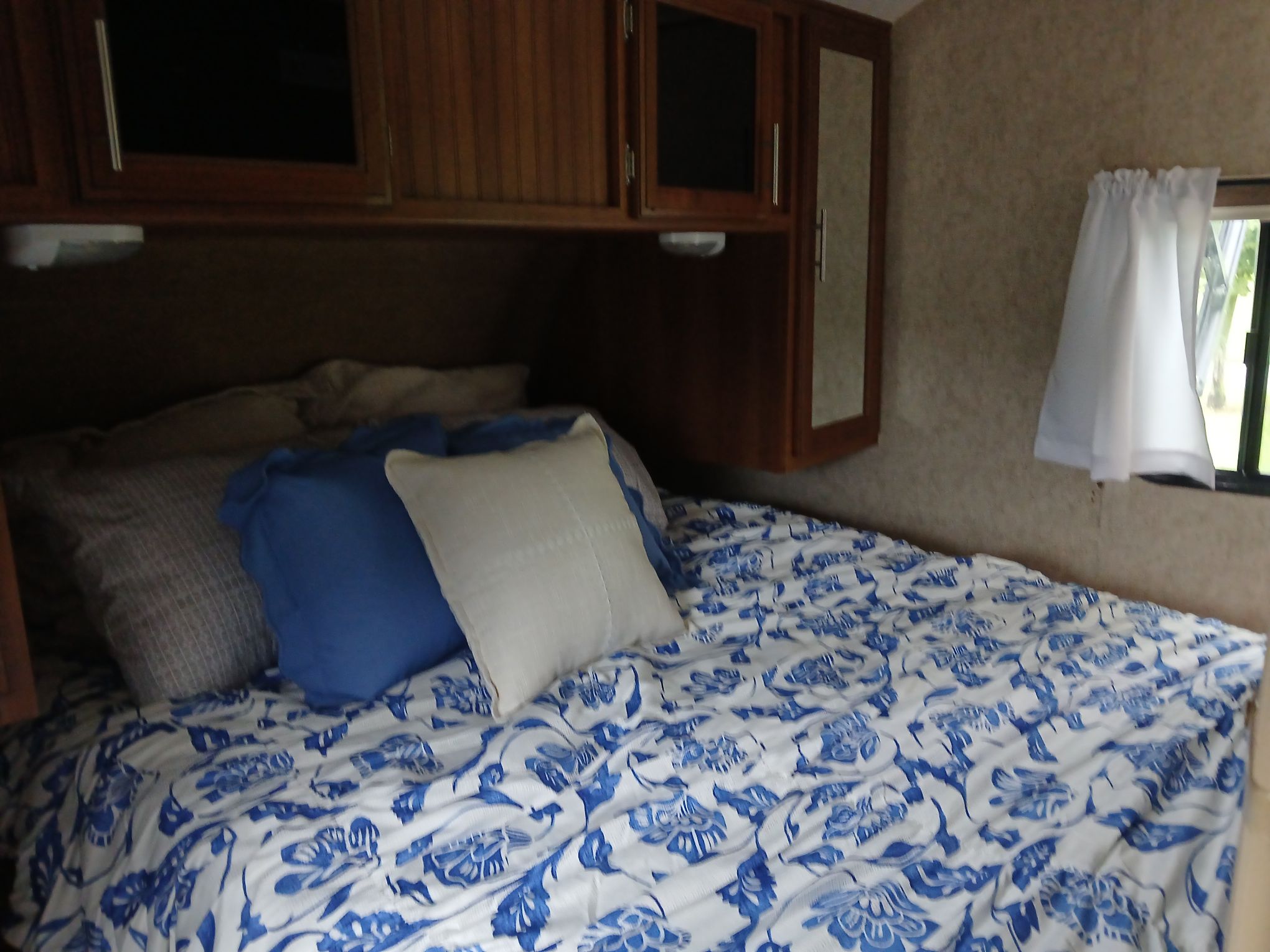 Inside an RV rental bedroom: A bed with blue and white floral bedding, various pillows, wooden overhead cabinets, and a window.. Forest River Tracer Air 2016