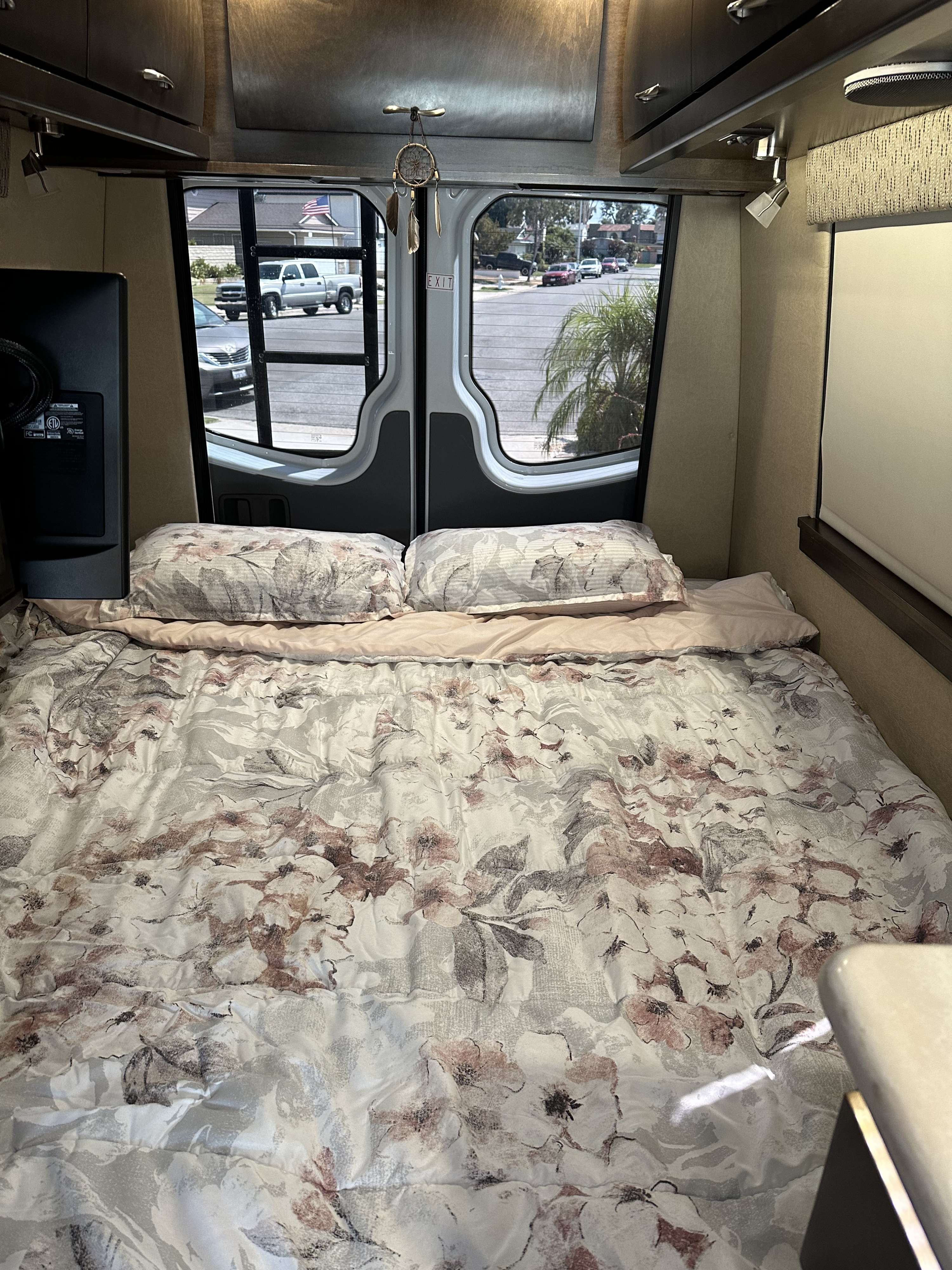 Inside an RV rental: a made bed with floral bedding, pillows, and a bright view of a residential street through the rear windows.. Mercedes-Benz Sprinter 2017
