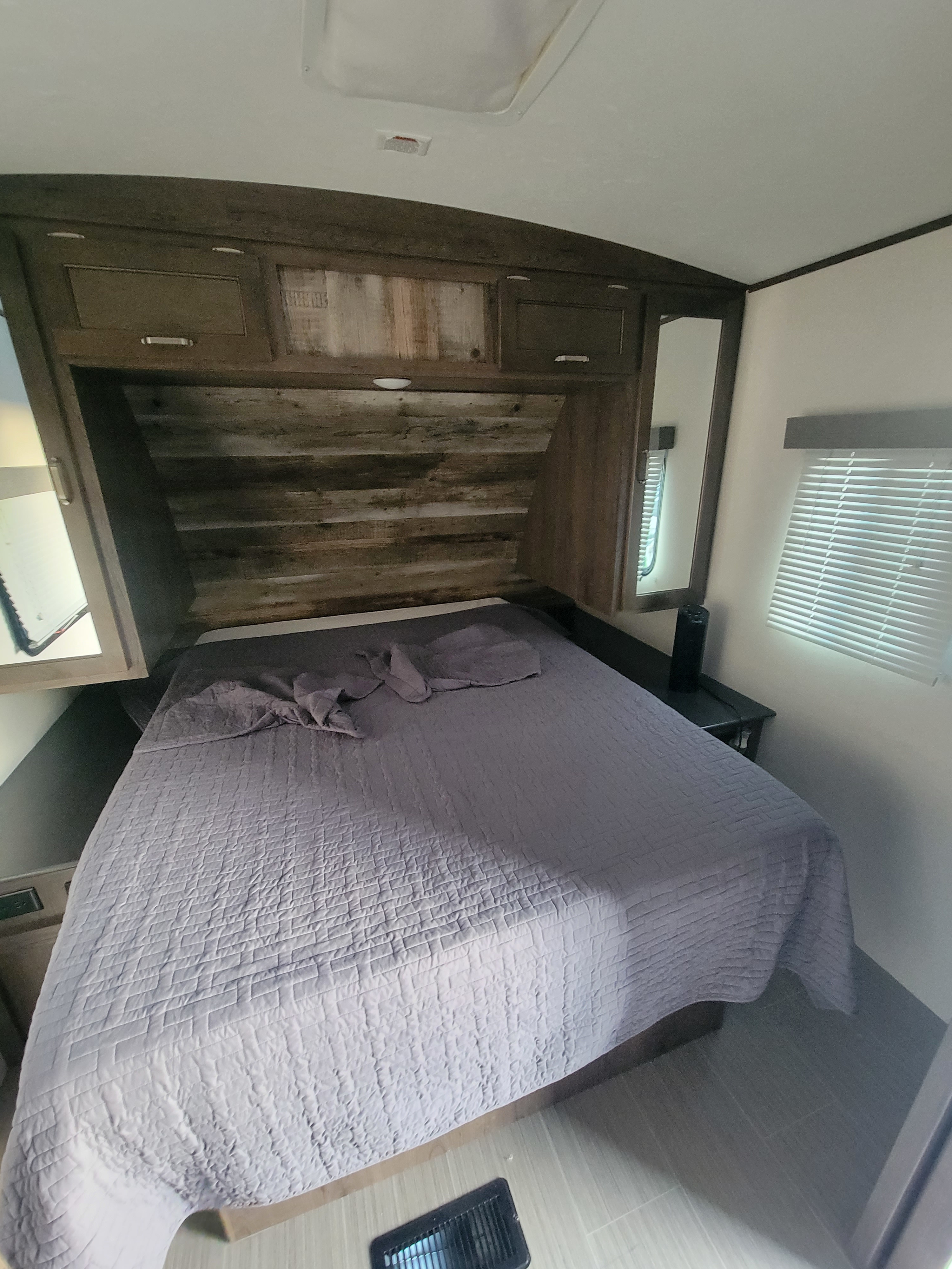 Comfortable RV rental bedroom with a grey quilted bed, wooden cabinets, side mirror, and window with blinds.. Keystone RV Passport GT 2022