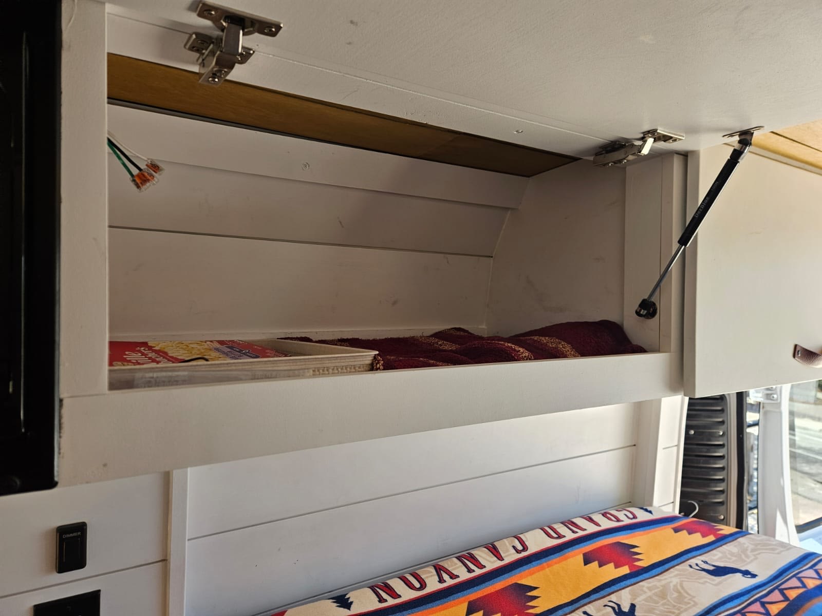 Inside an RV rental: open white overhead cabinet with blanket & food, plus a cozy bed with a patterned blanket below.. Dodge Ram 1500 2014