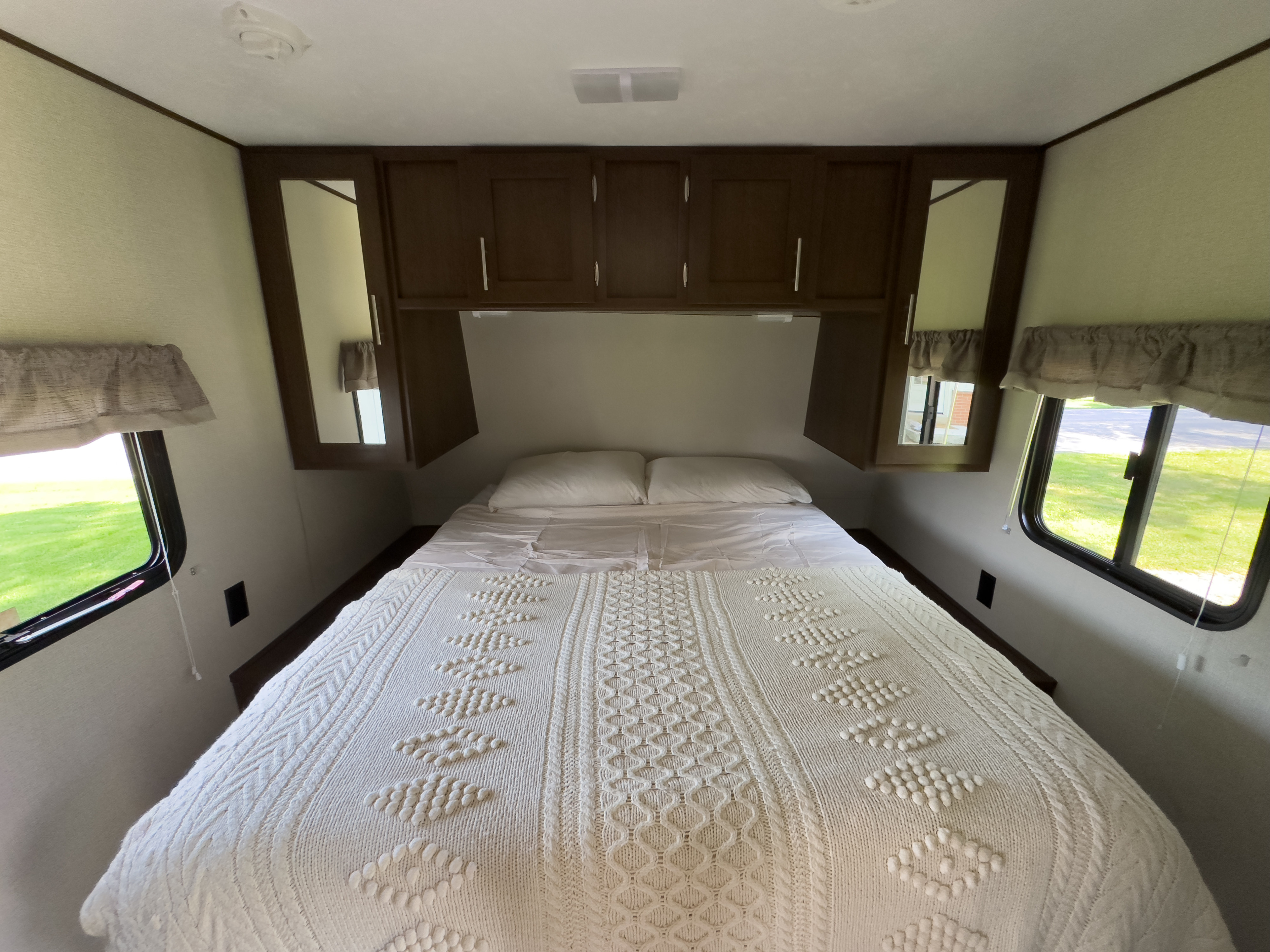 Cozy RV rental bedroom featuring a patterned white bed, overhead cabinets, and bright windows on both sides.. Avenger ATI 2019