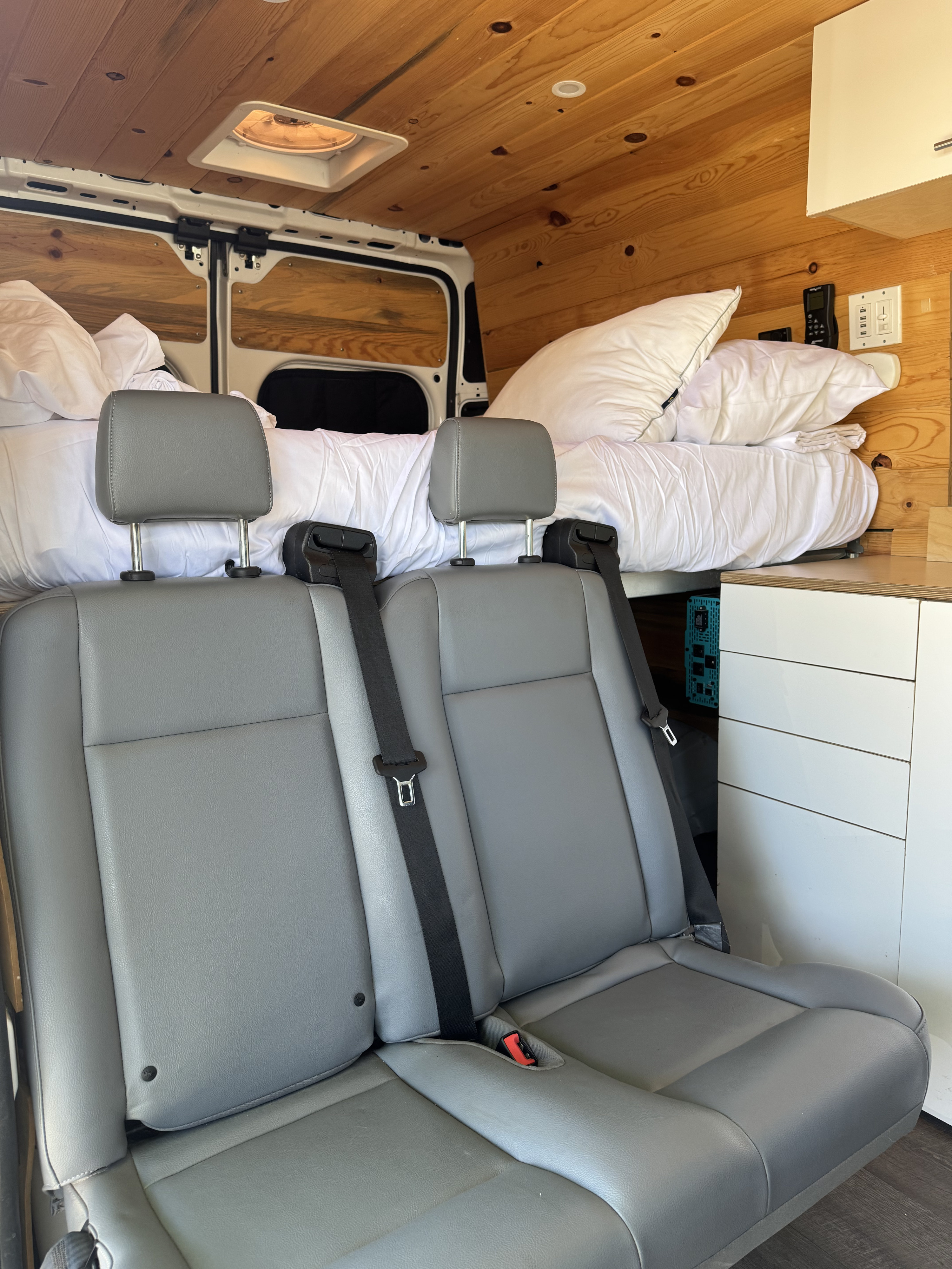 RV rental interior featuring grey passenger seating, a comfortable bed with white linens, wood paneling, and storage units.. Dodge B Van 2019