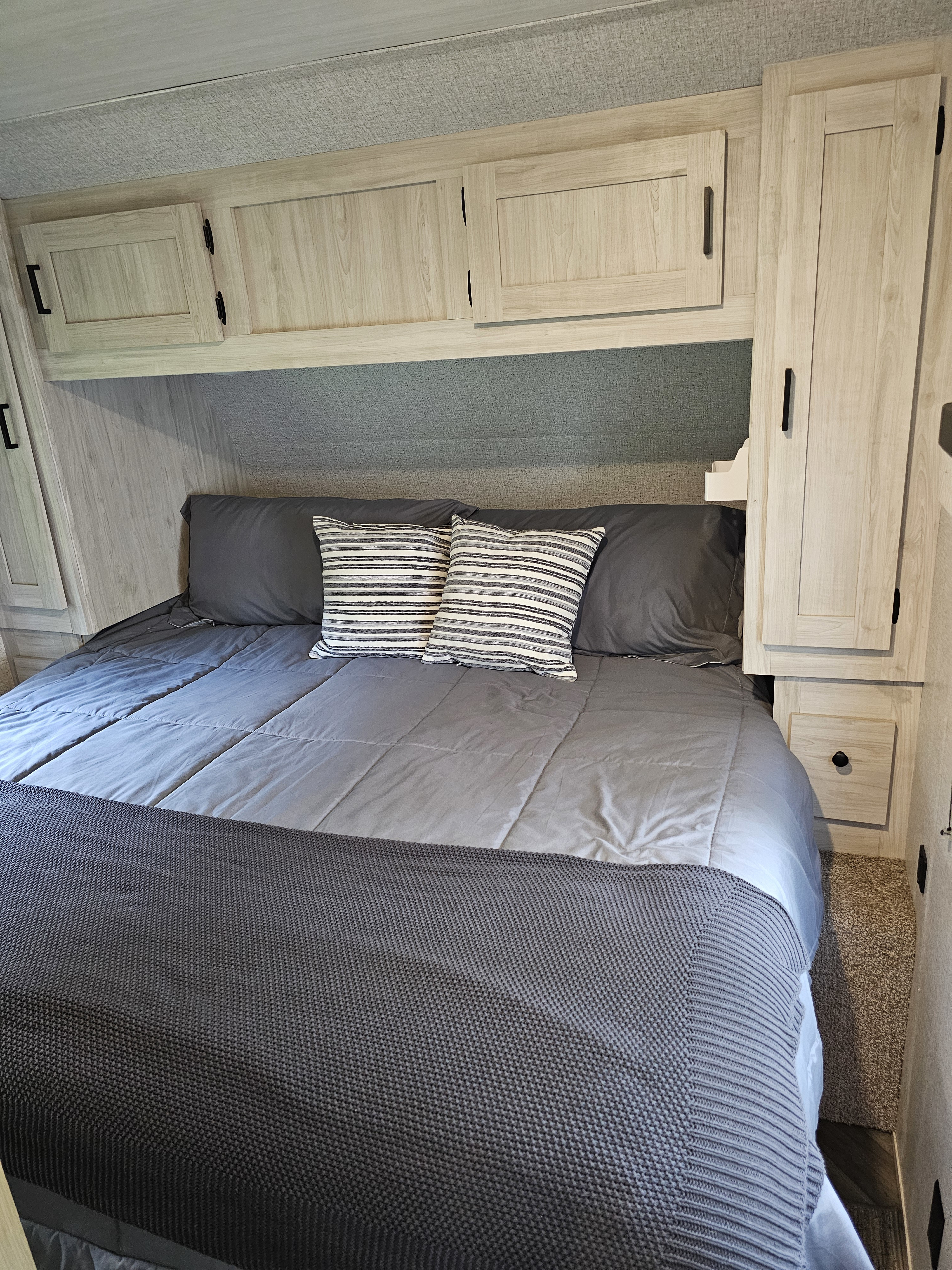 Cozy RV rental bedroom with light wood cabinets, gray bedding, and striped pillows.. East to West Della Terra 2022