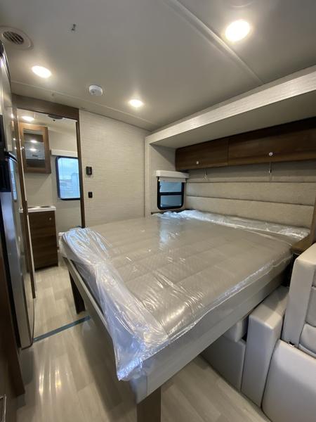 Experience comfort in this RV rental's cozy bedroom. Features a new bed with plastic wrap, overhead storage, and light wood floors.. Winnebago Navion 2023
