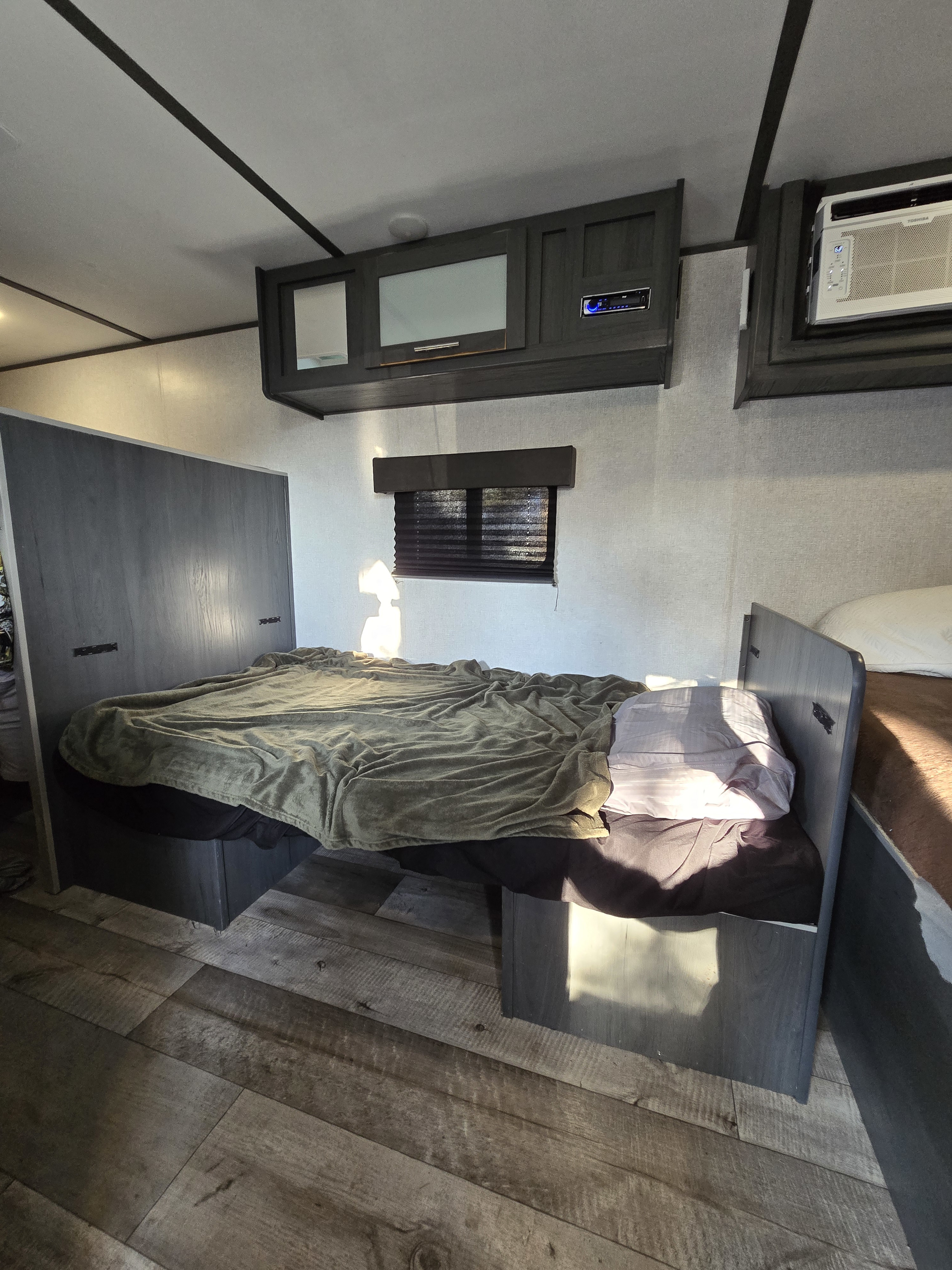 RV rental: Bright RV bedroom with compact bed, overhead cabinets with stereo, AC unit, and window with blinds. Wood floor.. Dutchmen ASPEN TRAIL 17BH 2021
