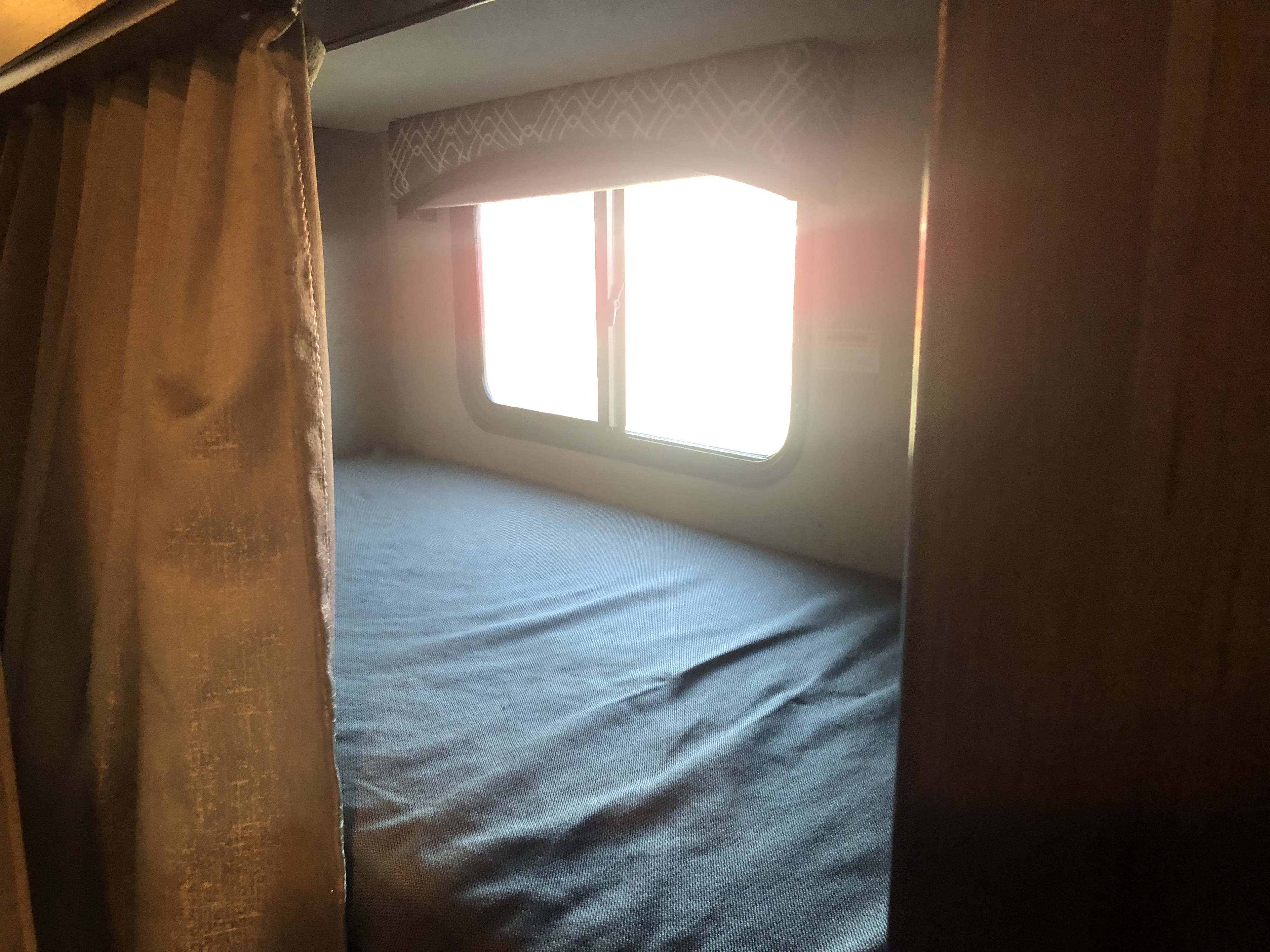 Inside an RV rental: a cozy bunk bed with a blue sheet, a bright window, and a brown privacy curtain.. Thor Chateau 2020