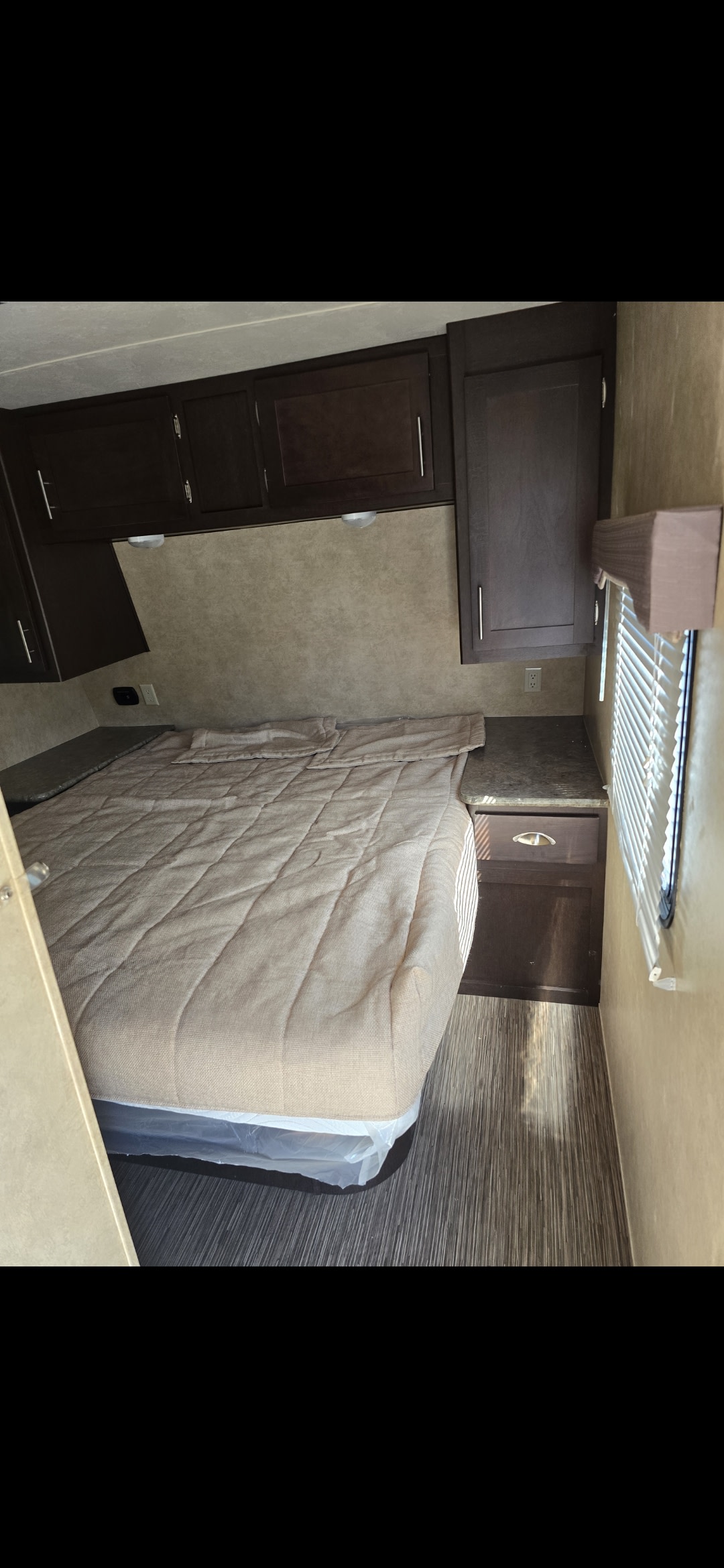 RV rental bedroom: quilted bed, dark wood overhead cabinets, and a nightstand with outlet by the window.. Forest River Evo series t2360 2015