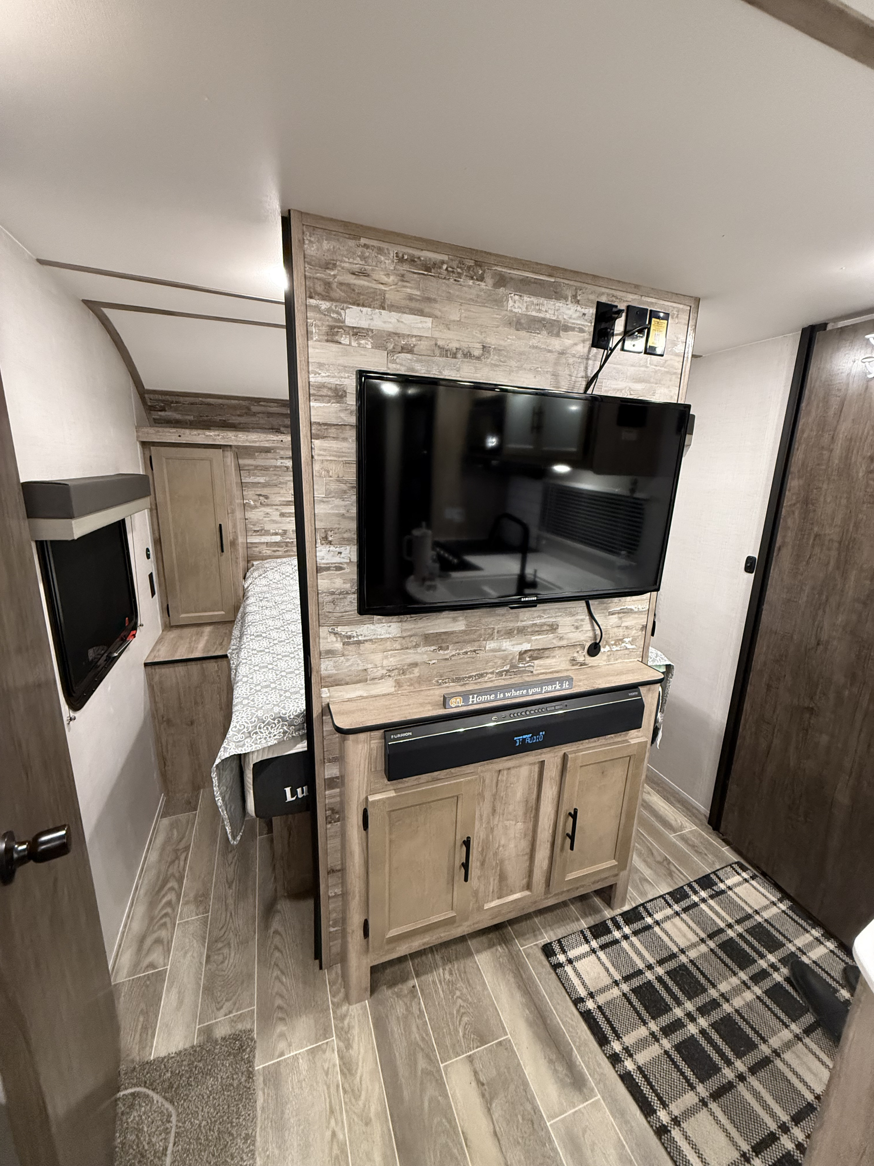 RV rental interior: media center with TV/soundbar, storage, bed, and wood-look floors. Kitchen visible in reflection.. Gulf Stream Ameri-Lite 2023