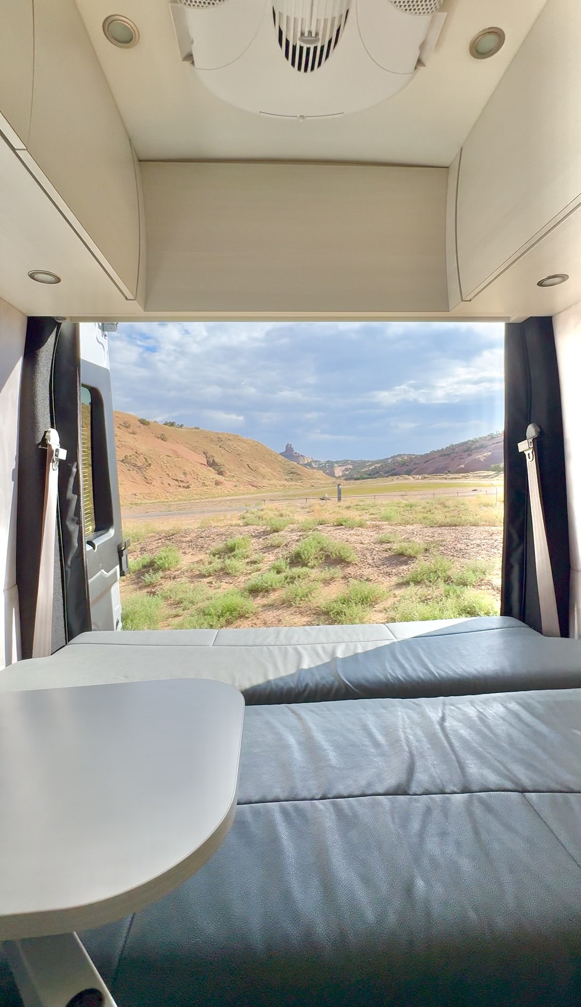 RV rental interior view: a cozy bed & table frame a scenic desert landscape with hills and road.. Pleasure-Way Pleasure-Way Class B 2023