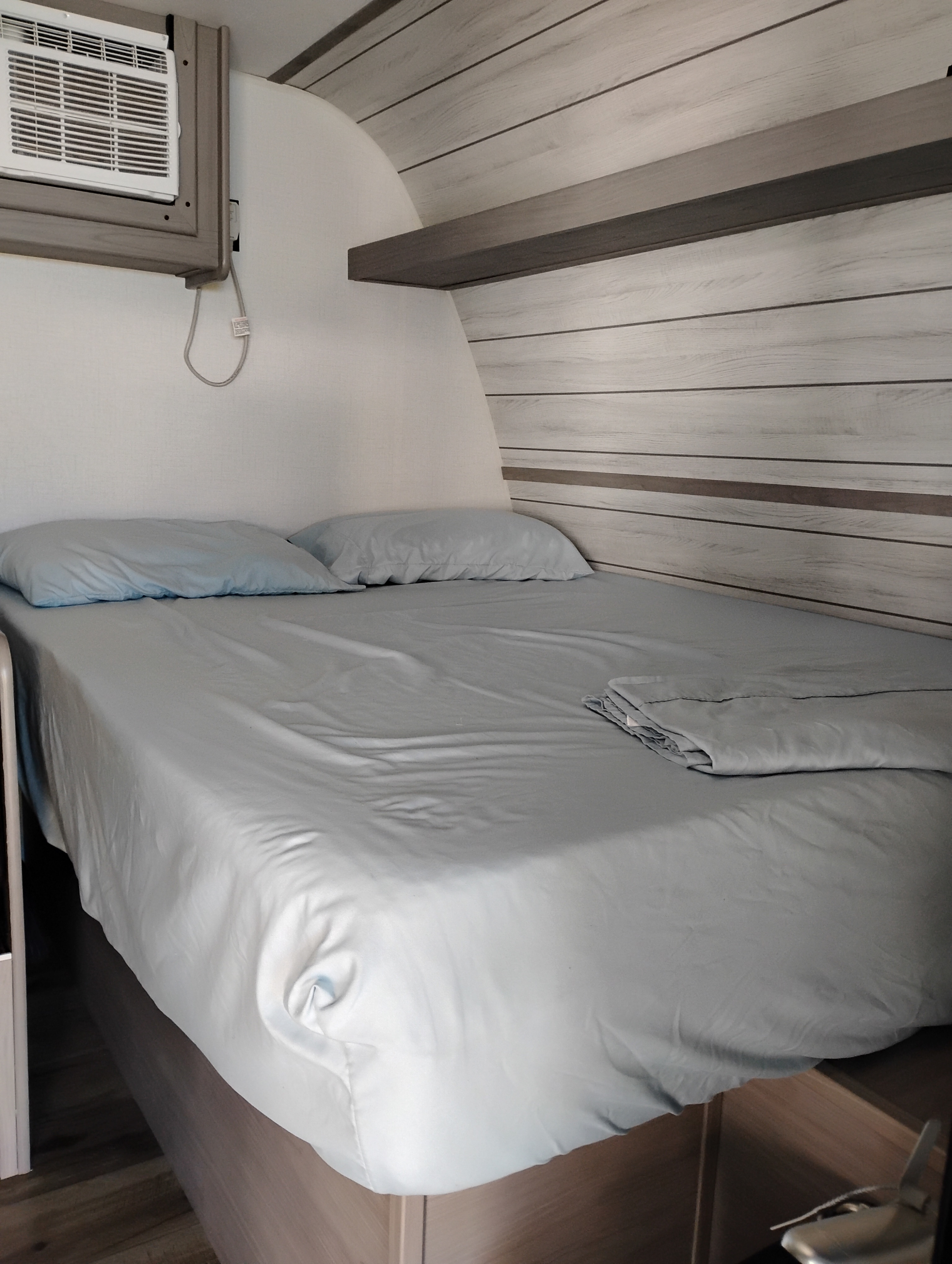 Inside an RV rental, a compact and cozy bedroom features a bed with light blue linens, a mounted AC unit, and wood-paneled walls.. Dutchmen Coleman 17B 2024