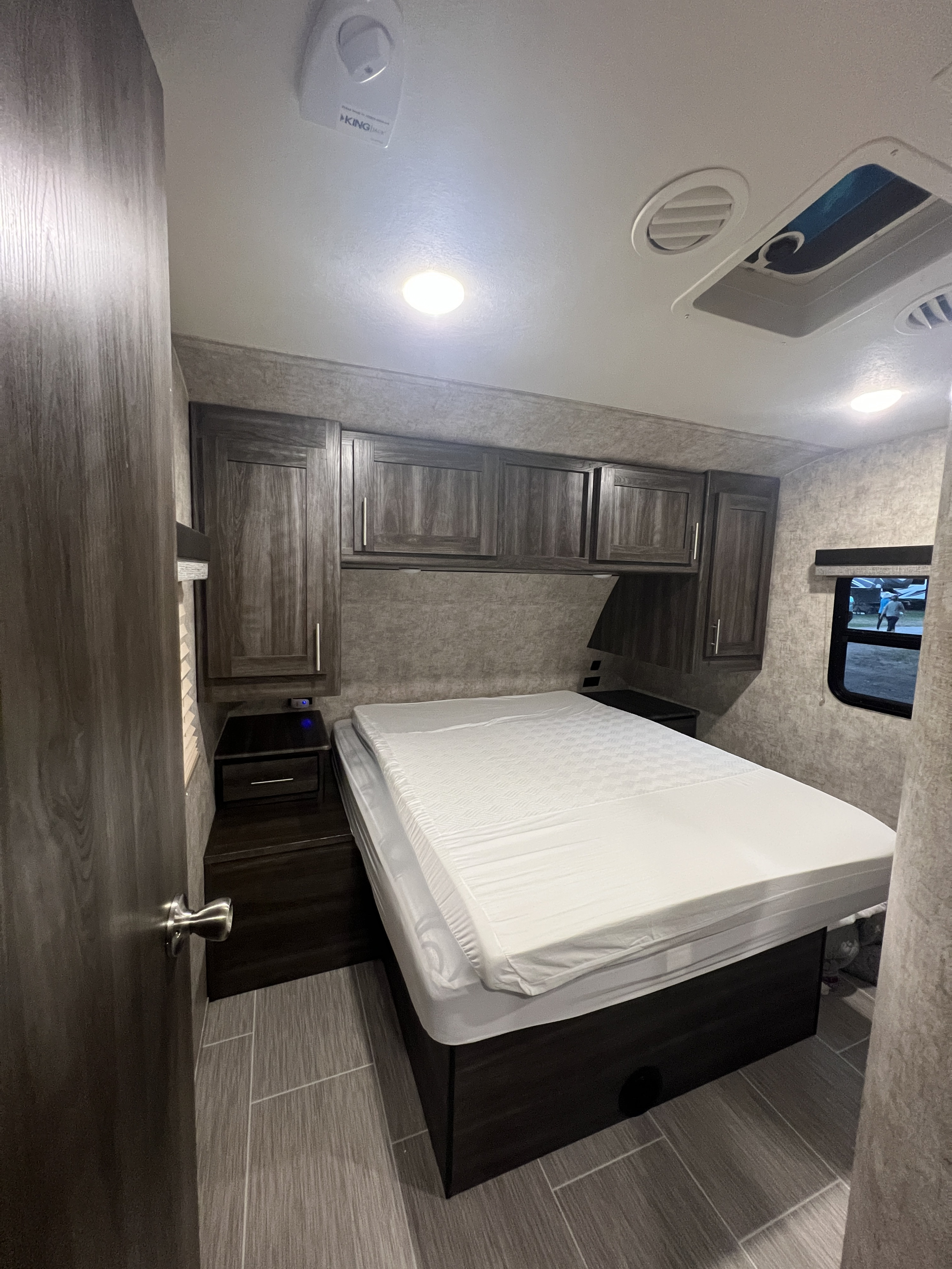 Compact RV rental bedroom with a comfy bed, dark wood cabinets, bright lighting, and a side window.. Jayco highland Ridge 2019