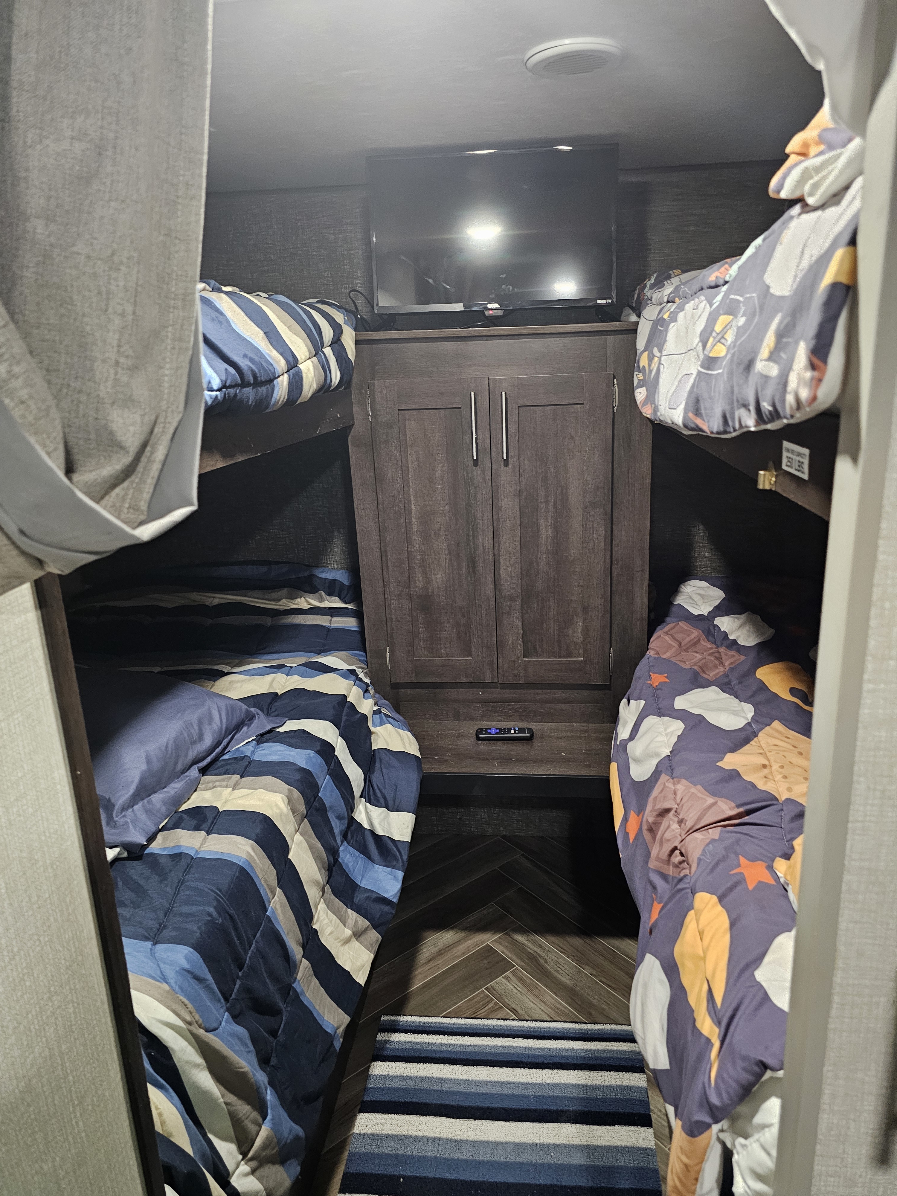 Inside an RV rental, showing compact bunk beds with striped and s'mores bedding, central TV, and cabinet storage.. KZ Sportsman 2021
