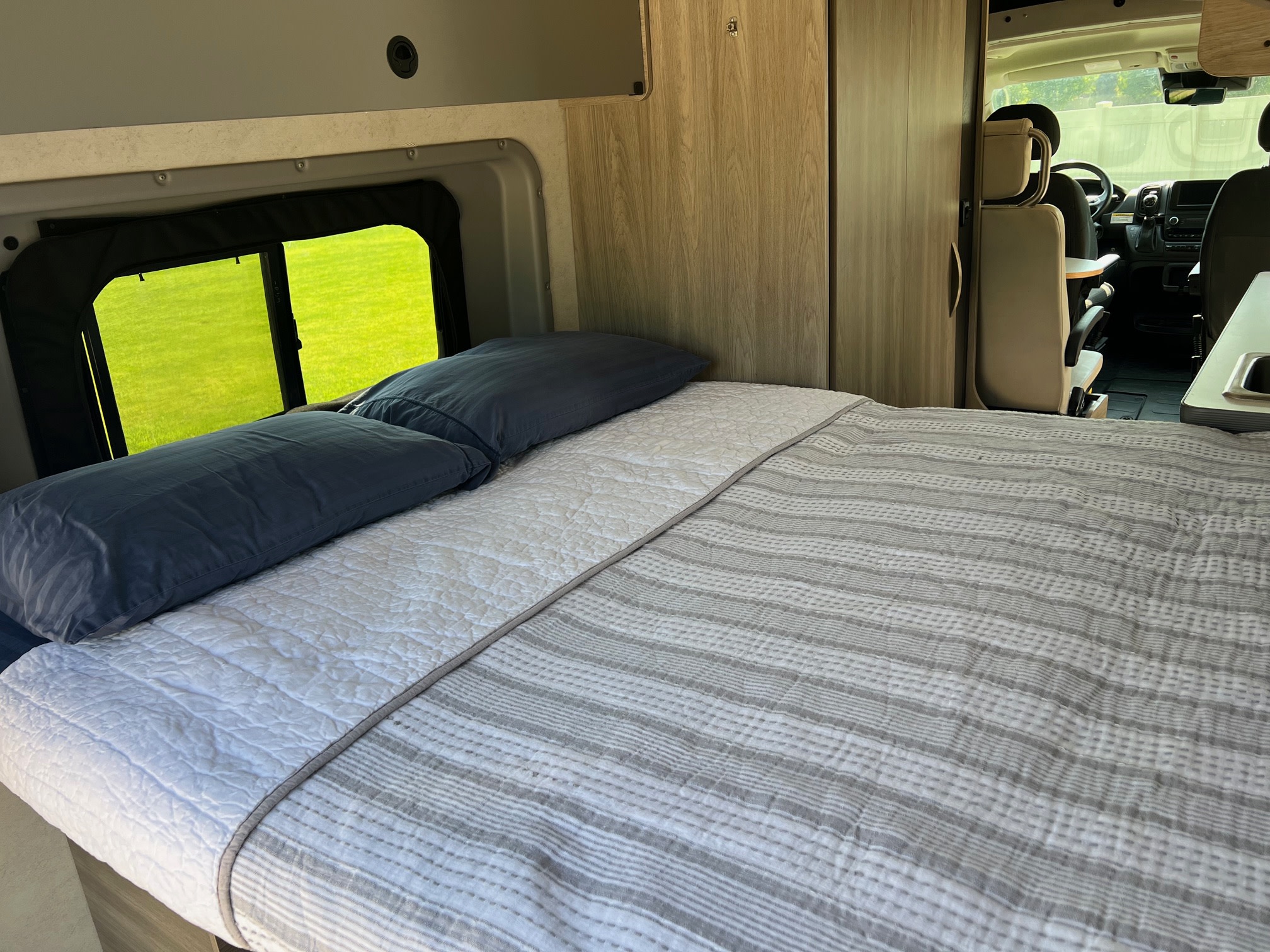 Inside an rv rental: a cozy bed with pillows & quilt, a bright window to grass, and the front camper cabin.. Winnebago Solis 2023