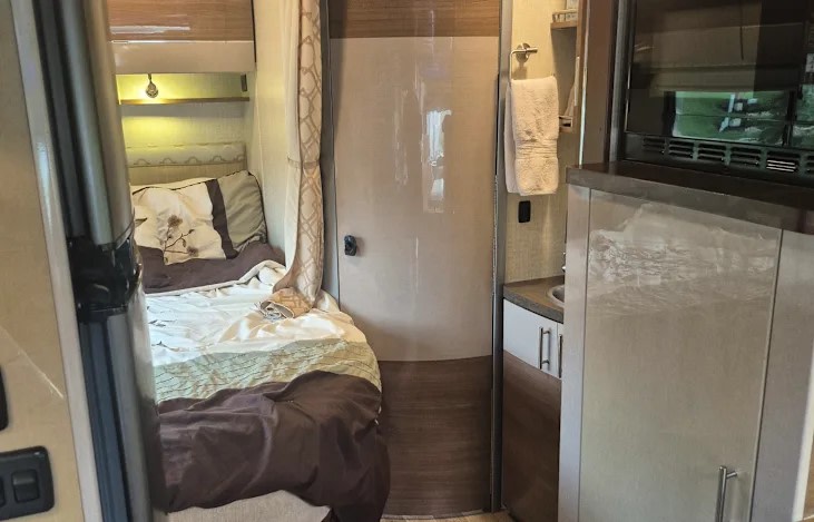RV rental interior: A bedroom with patterned bedding, a bathroom door, and a compact kitchenette with microwave.. Winnebago View 2016