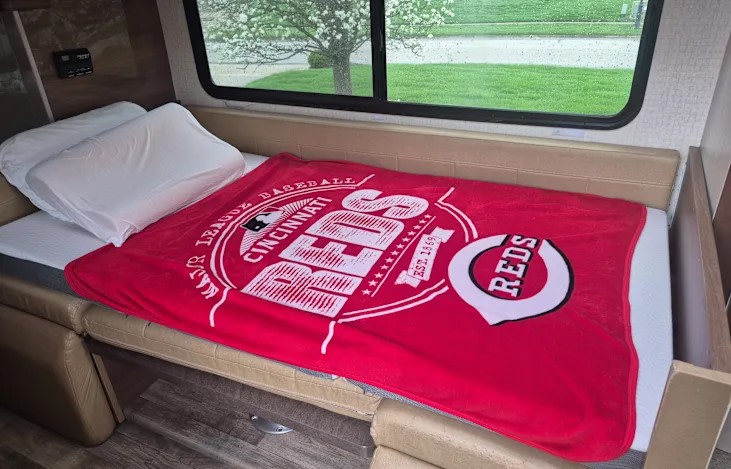 Cozy RV rental bed with white pillows and a Cincinnati Reds blanket. A comfortable sleeping area with a window view of greenery.. Winnebago View 2016