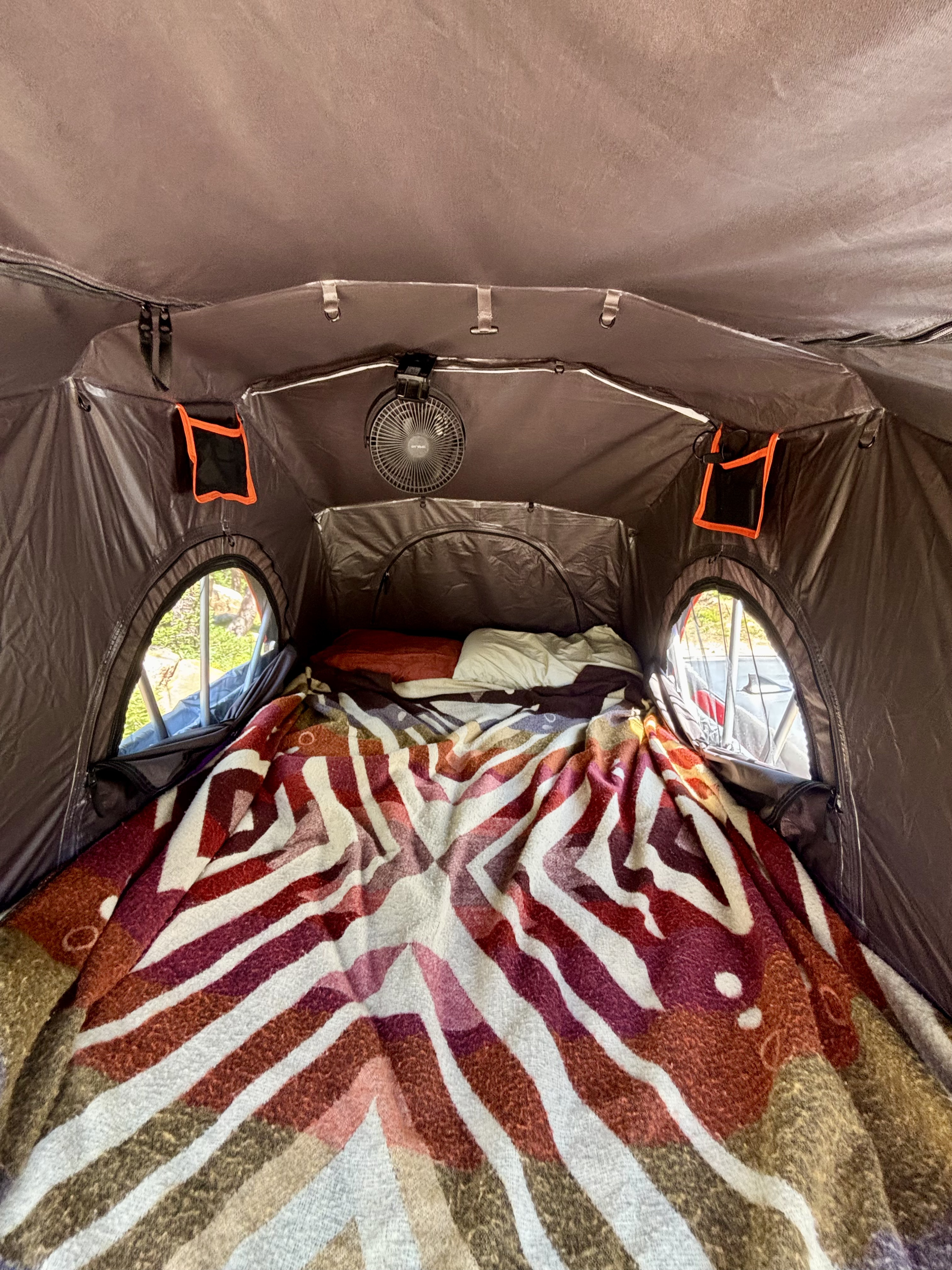 Interior of an RV rental showing a cozy bed with a colorful blanket, pillows, fan, and oval windows.. Toyota Tacoma 2012