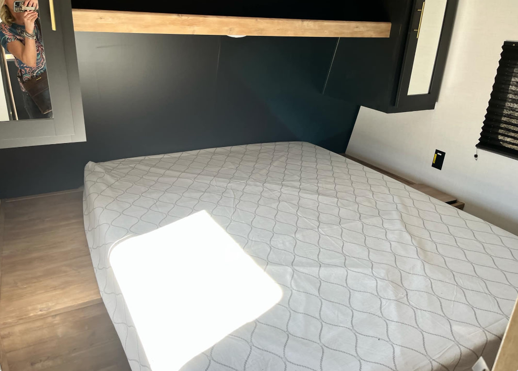Compact RV rental bedroom with a comfy bed, dark accent wall, and wood shelf. A person is reflected in the mirror.. Keystone RV Springdale 2023