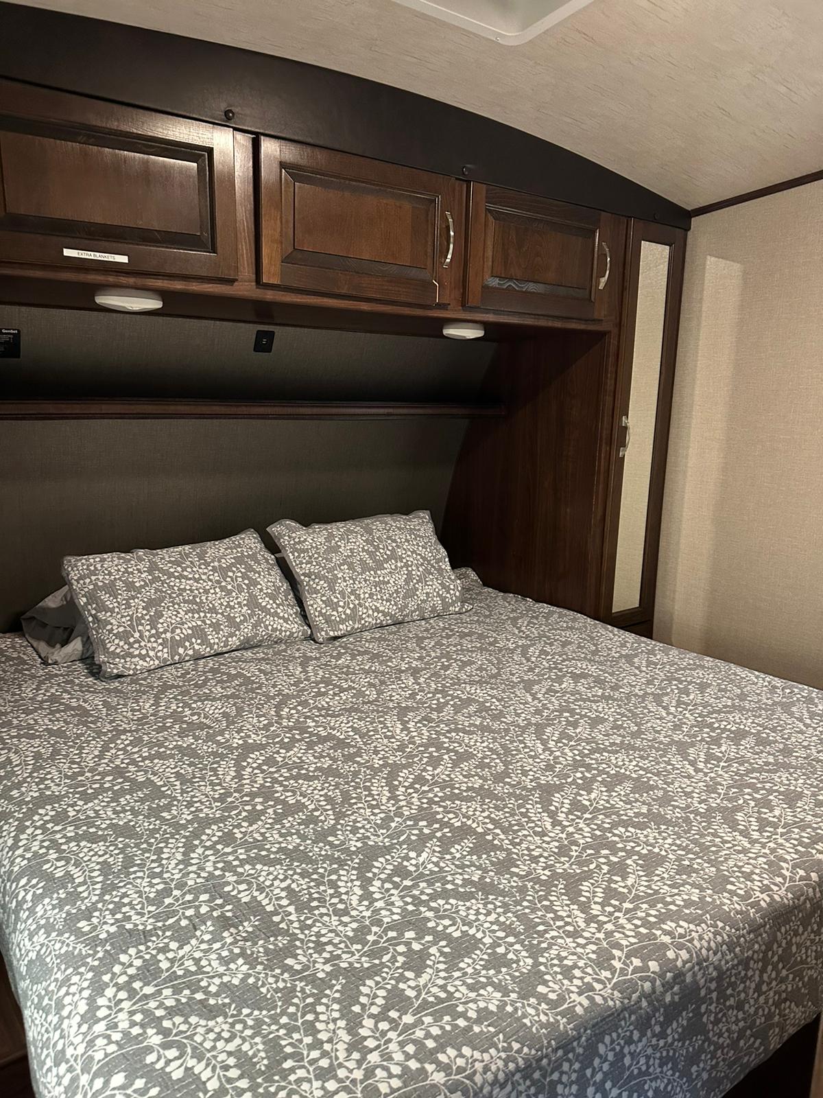 Cozy RV rental bedroom featuring a comfy bed with patterned linens, overhead storage cabinets, and a side mirror.. Heartland Torque 2018