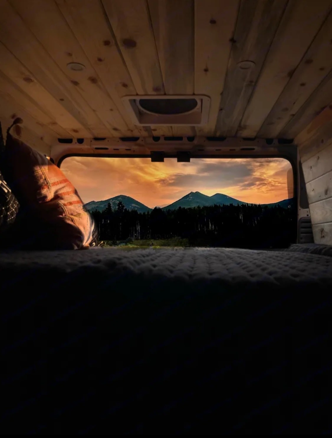 RV rental: Inside a cozy, wood-paneled camper van, a bed faces a stunning sunset over silhouetted mountains and trees.. Dodge Ram Promaster 2500 2021