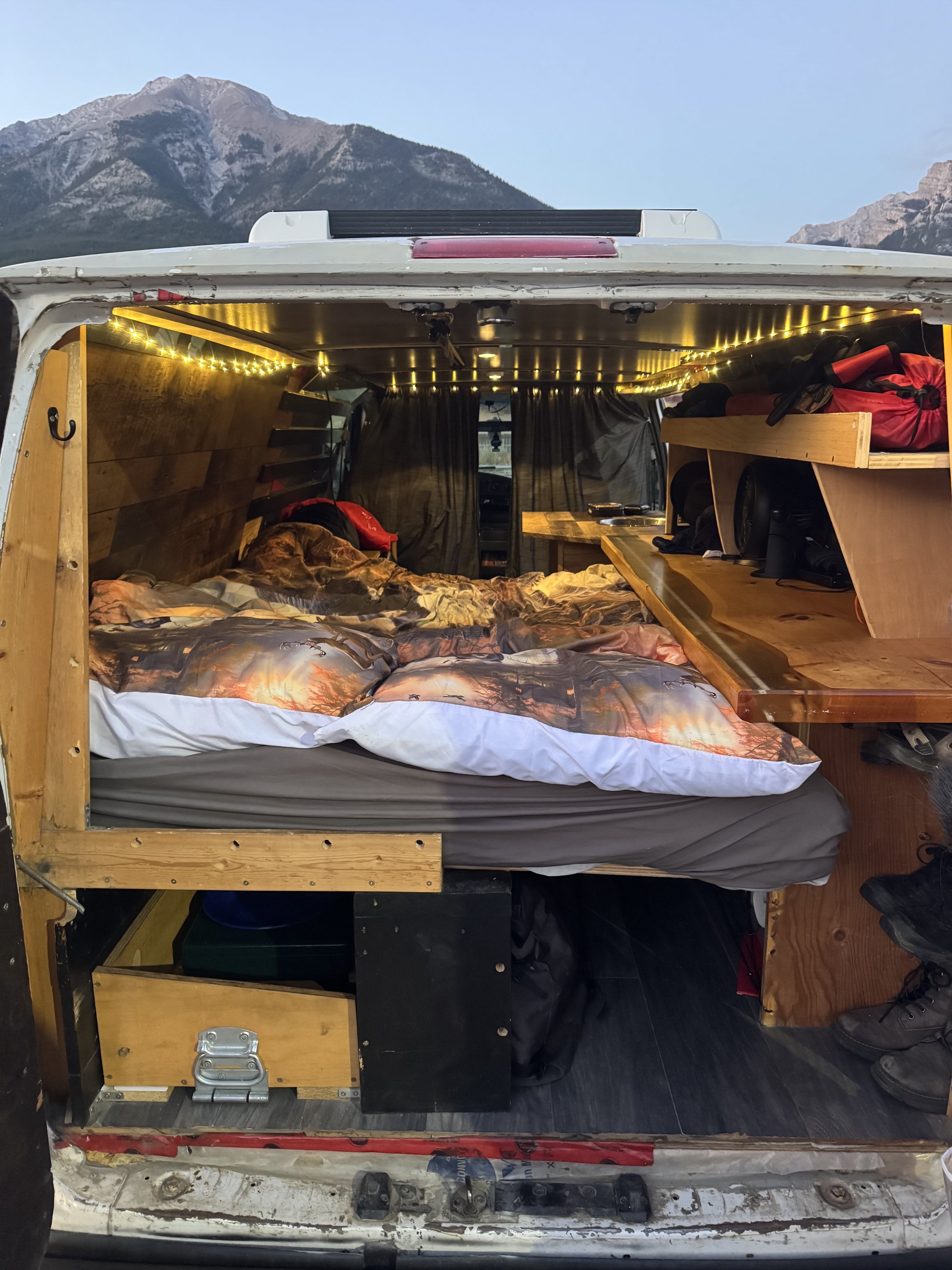 View from inside a converted RV rental: a bed with patterned duvet, wooden interior, and mountains outside.. Ford E350 Box Van 2006