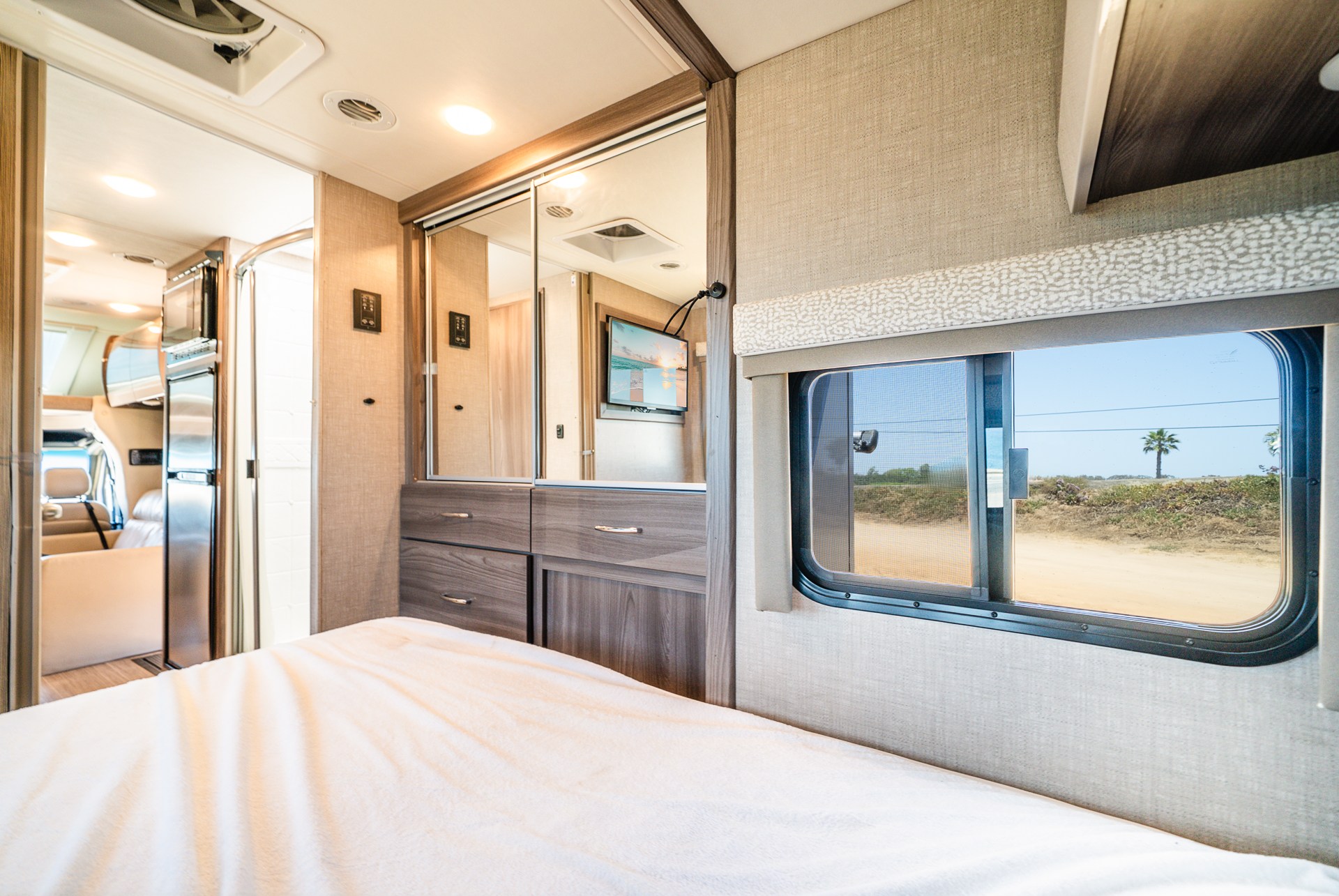 Cozy rv rental bedroom with a white bed, mirrored cabinets, and a window looking out to a desert landscape with a palm tree.. Thor Motor Coach Delano SV 2023