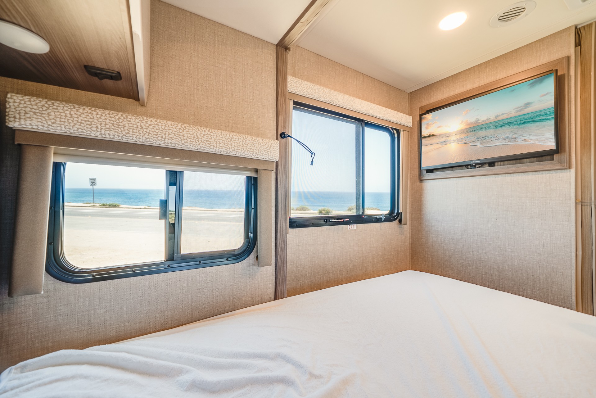 Cozy RV rental bedroom with a stunning ocean view from multiple windows, featuring a comfy bed and wall-mounted TV.. Thor Motor Coach Delano SV 2023