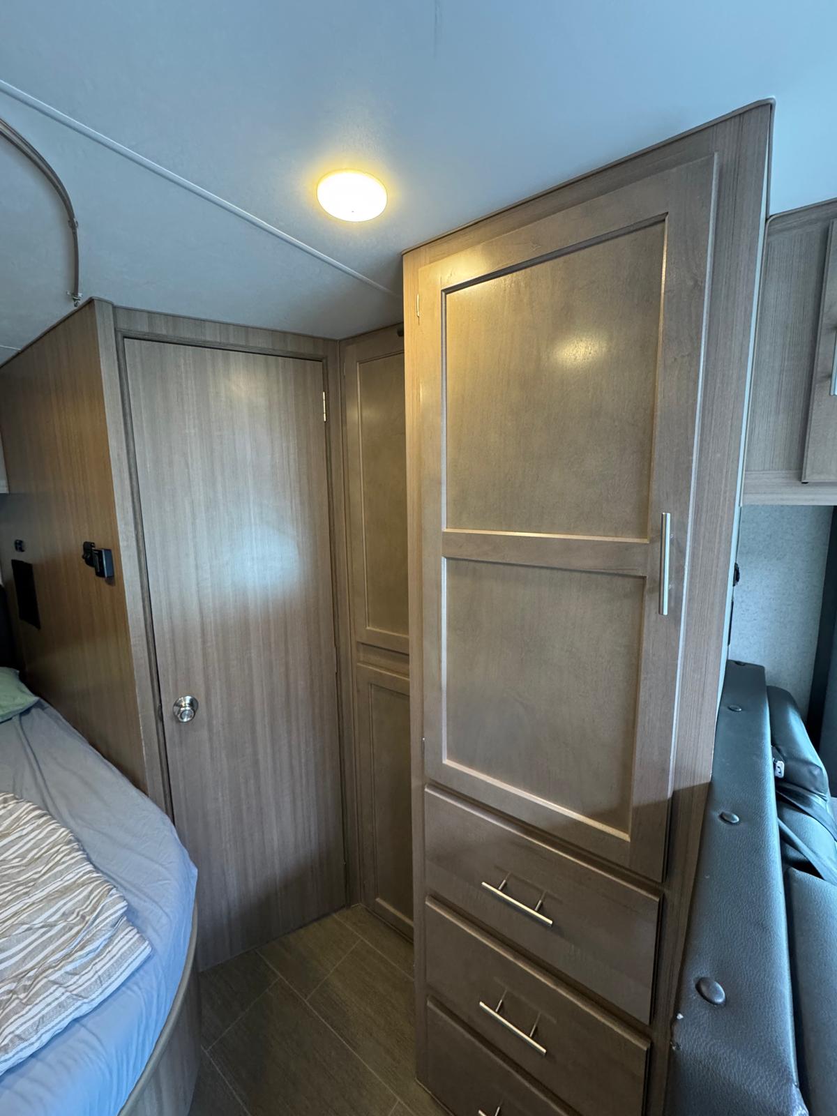 Inside a modern RV rental: wood-paneled interior with a bed, closed door, and tall storage cabinet with drawers.. Ford Adventurer 2021