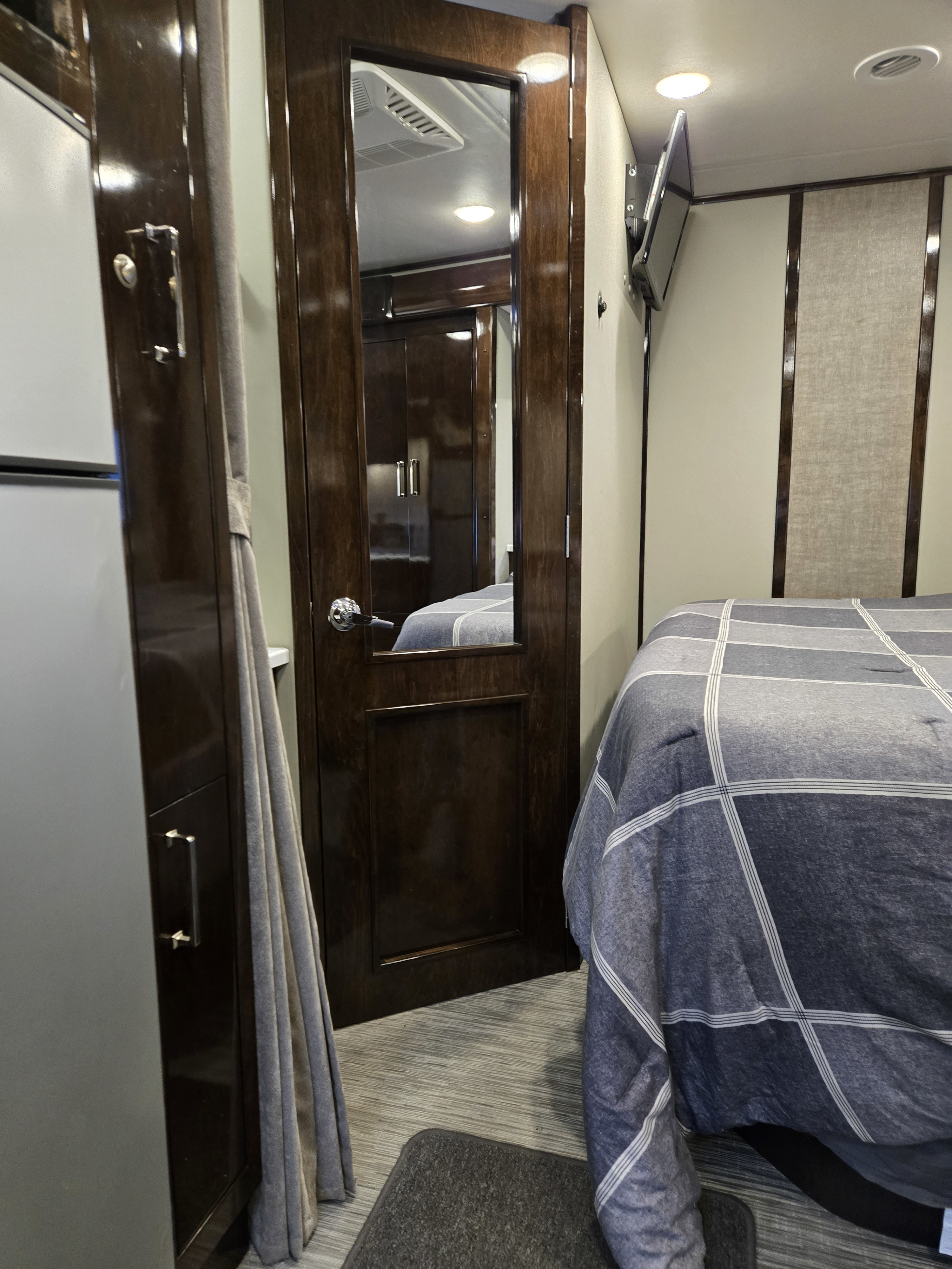 RV rental bedroom interior featuring a compact bed with plaid bedding, mirrored door, and wall-. Renegade Custom Coaches Vienna 2021