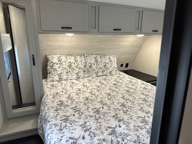Compact RV rental bedroom with a bed featuring black and white floral bedding, overhead cabinets, and a small nightstand.. Forest River Vibe 2023
