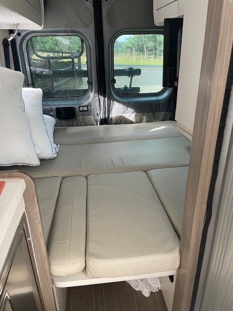 Interior view of an RV rental, featuring a comfortable bed setup with light cushions, rear windows, and a wall-mounted TV.. Thor Tellaro 2022