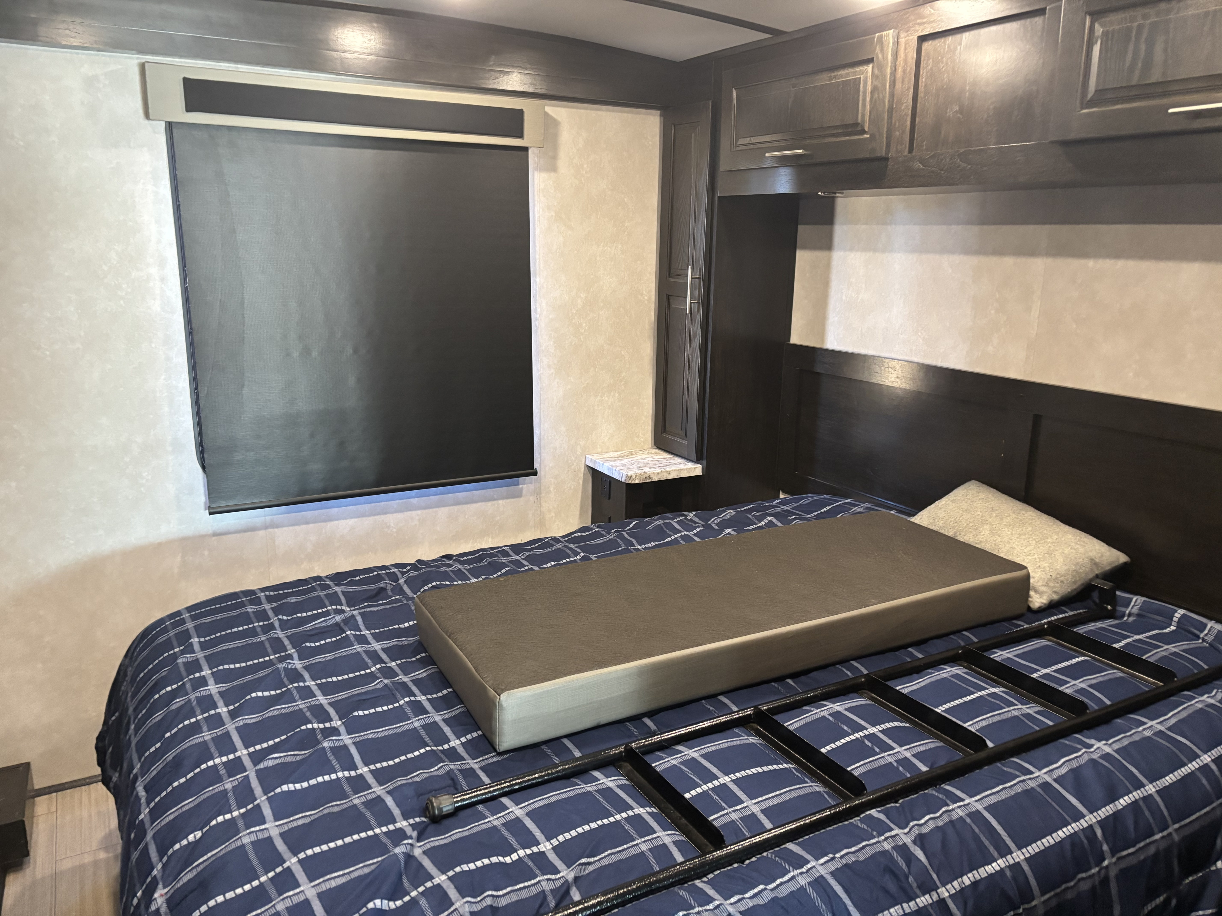 This RV rental bedroom offers dark wood cabinetry, a bed with blue plaid, and a window with a blackout shade. A ladder rests on the bed.. FR3 FR3 Motorhome 2021