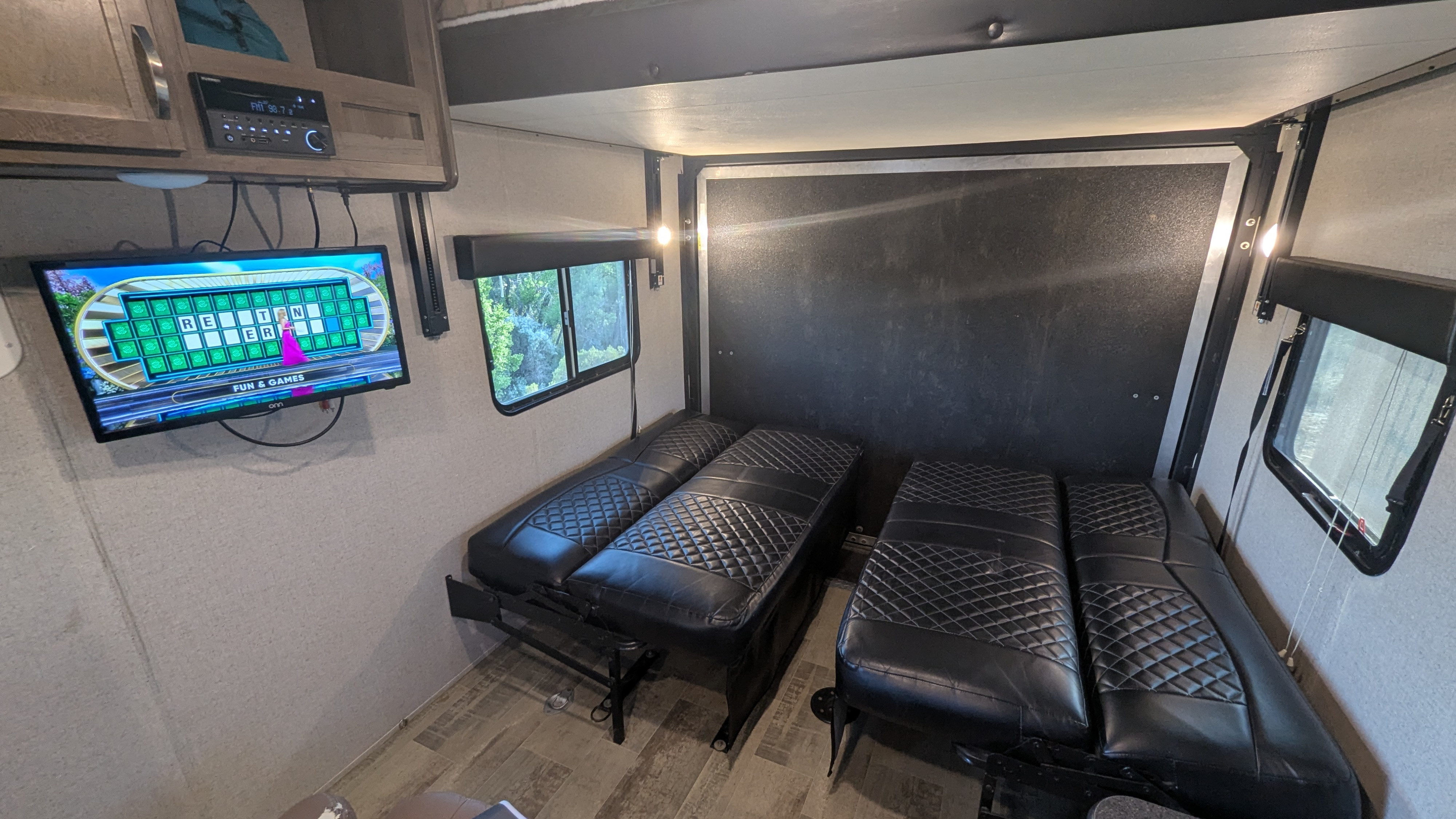 An RV rental interior showing a TV playing Wheel of Fortune, windows, and convertible black quilted beds.. Dutchmen Rubicon 2018